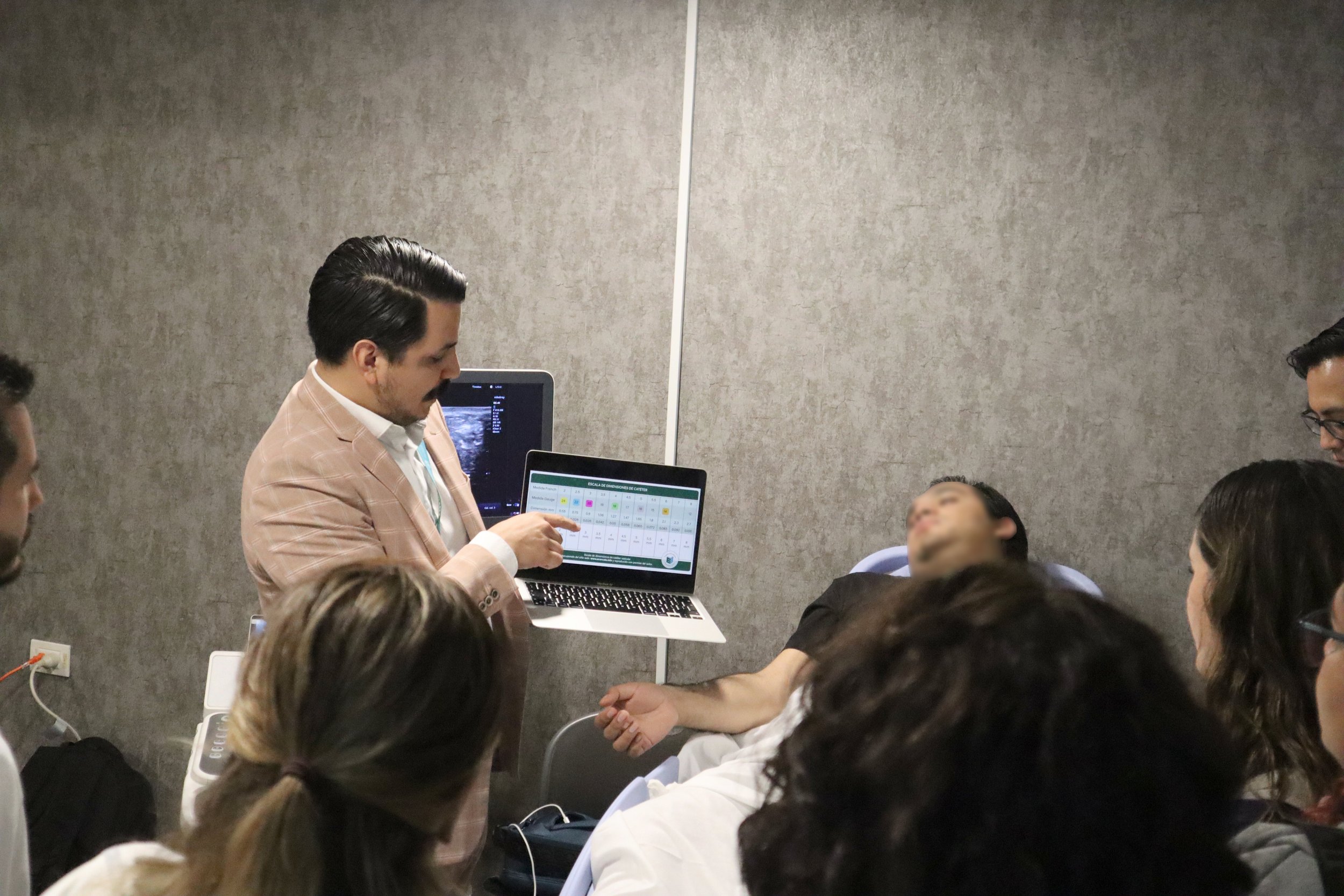 Un médico explica resultados de un ultrasonido a un paciente en una consulta médica, rodeado de varias personas en una sala de consulta. ACÉRCATE Asociación para certificación y capacitación Técnica Catéter PICC