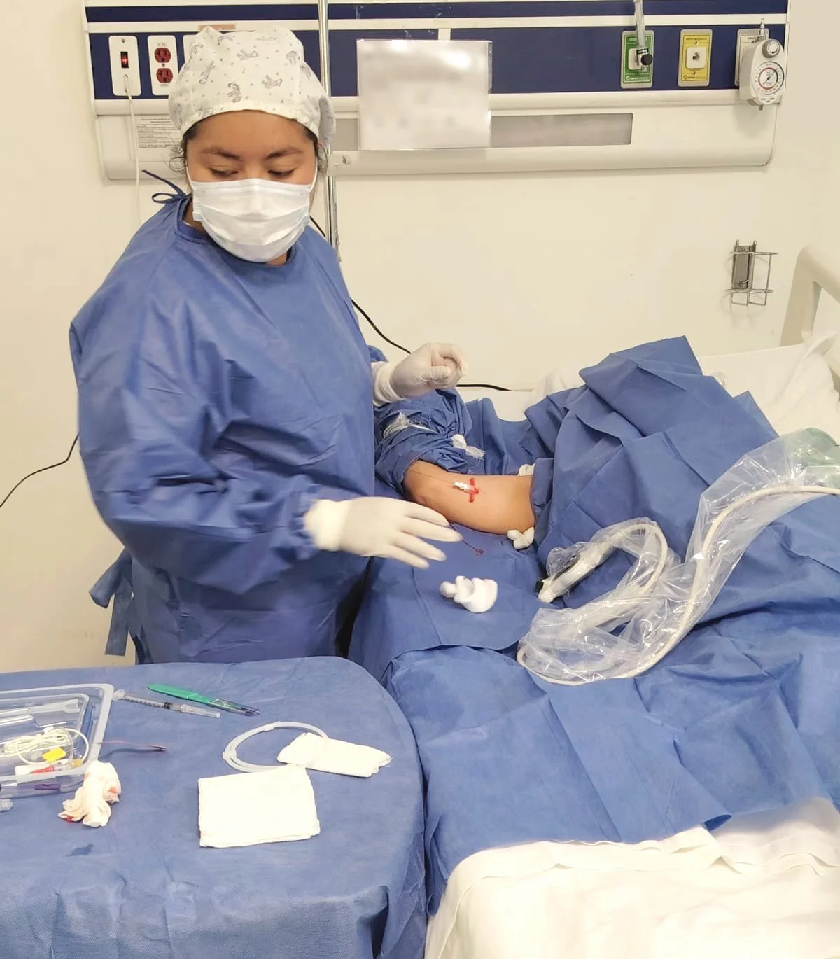 Enfermera en una sala de operaciones realizando un procedimiento médico en el brazo de un paciente. La enfermera usa guantes, mascarilla y bata médica. En la mesa de alrededor, hay instrumentos quirúrgicos y vendajes. ACÉRCATE para PICC