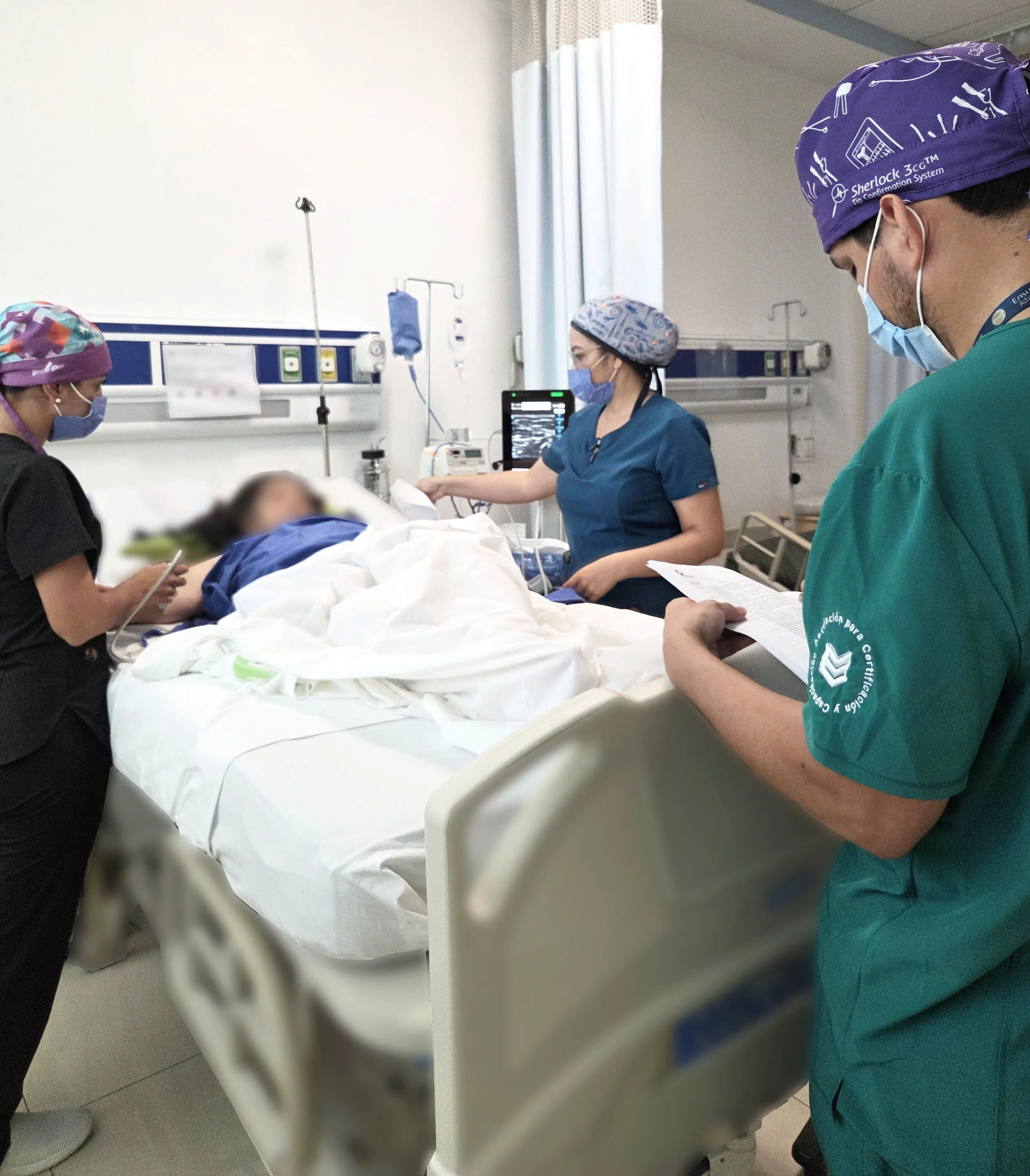 Personal médico atiende a una paciente en una cama de hospital, mientras otros profesionales revisan y documentan información en la sala de urgencias o cuidados intensivos, con monitores y equipo médico en el fondo. ACÉRCATE Catéter PICC