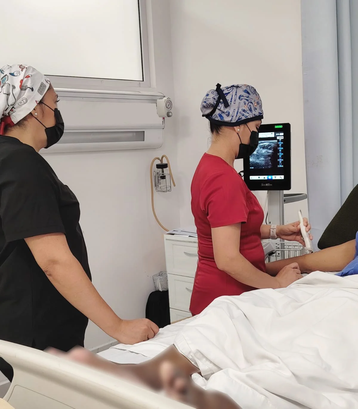 Dos profesionales de la salud realizan un ultrasonido en un paciente en una sala de hospital. Ambas llevan cubrebocas y gorros médicos, uno con uniforme negro y otro con uniforme rojo. ACÉRCATE Catéter PICC