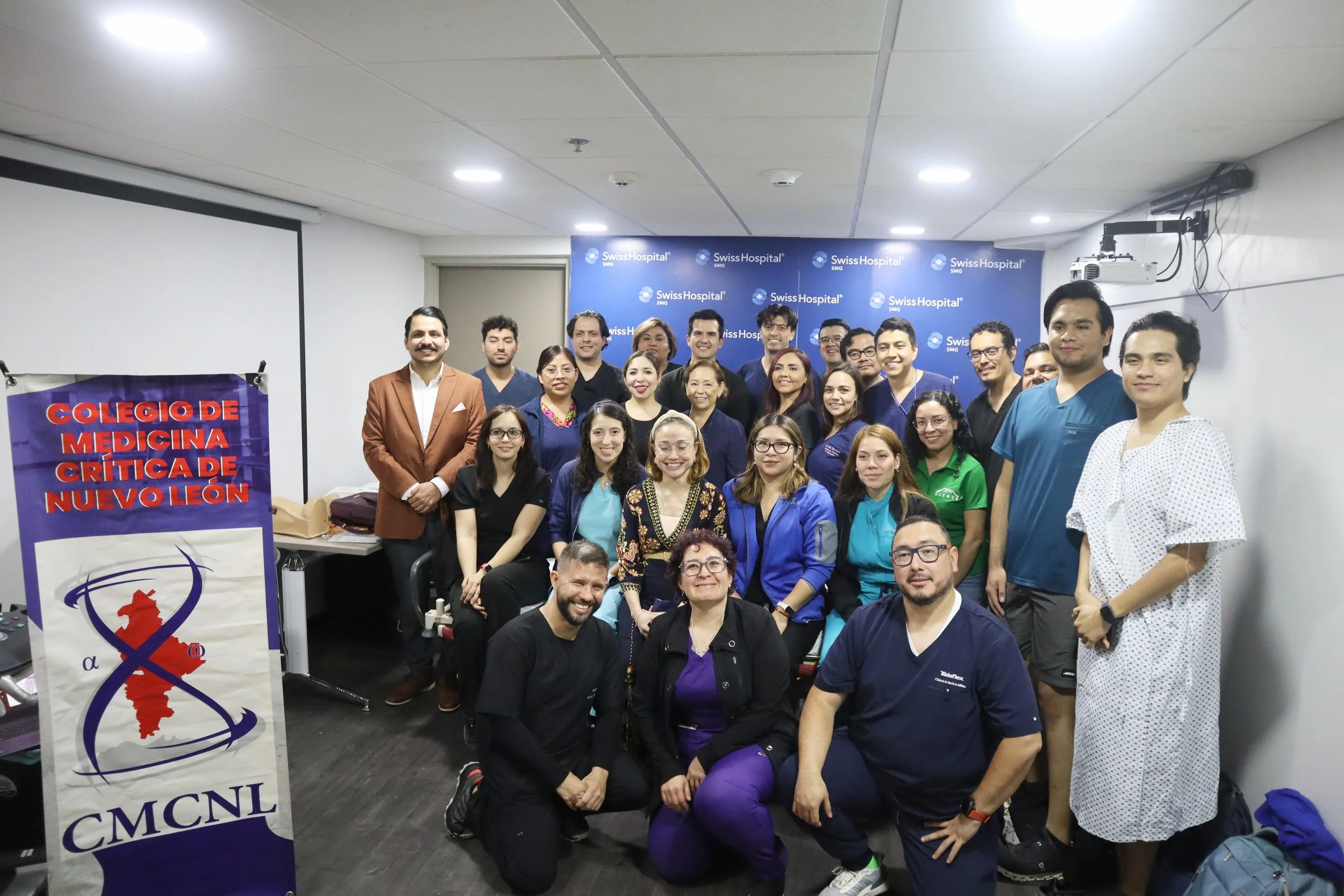 Grupo de personas en una clínica médica en un salón con fondo azul que dice 'Swiss Hospital'. Algunos llevan uniforme de médicos y otros ropa casual o de oficina, posando para una foto grupal. ACÉRCATE Asociación para certificación y capacitación Téc