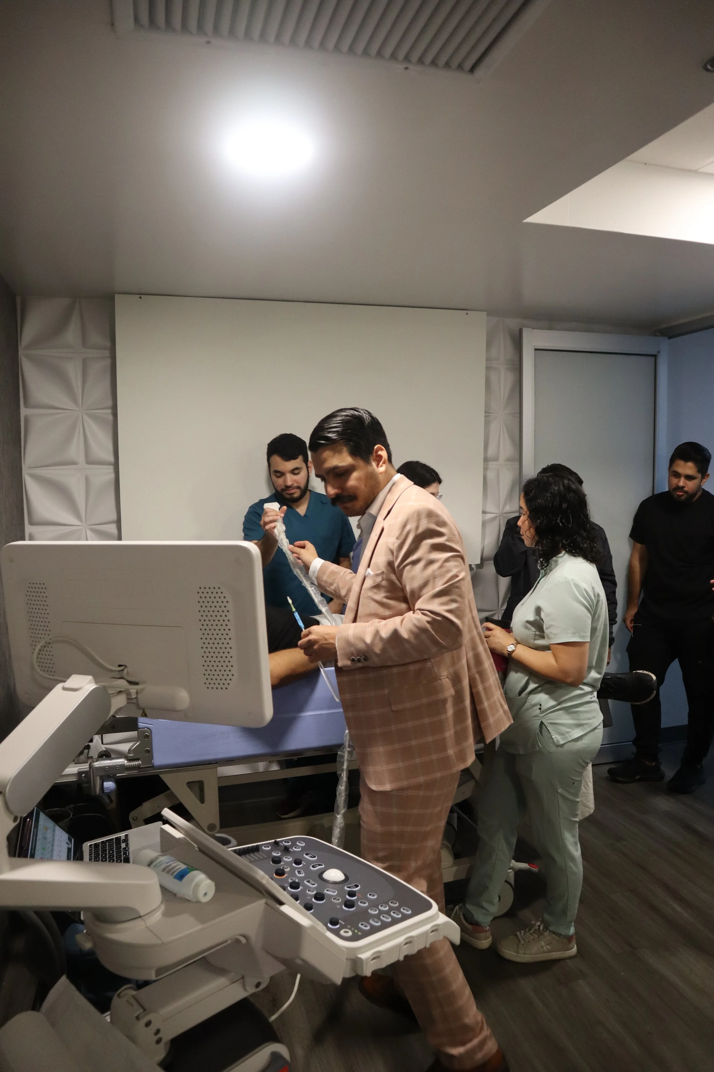 Un grupo de personas en una sala de parto o emergencia médica, incluyendo un hombre con un traje de cuadros en el centro, atendiendo a una paciente en la cama del hospital, rodeados de otros profesionales de la salud. ACÉRCATE para PICC