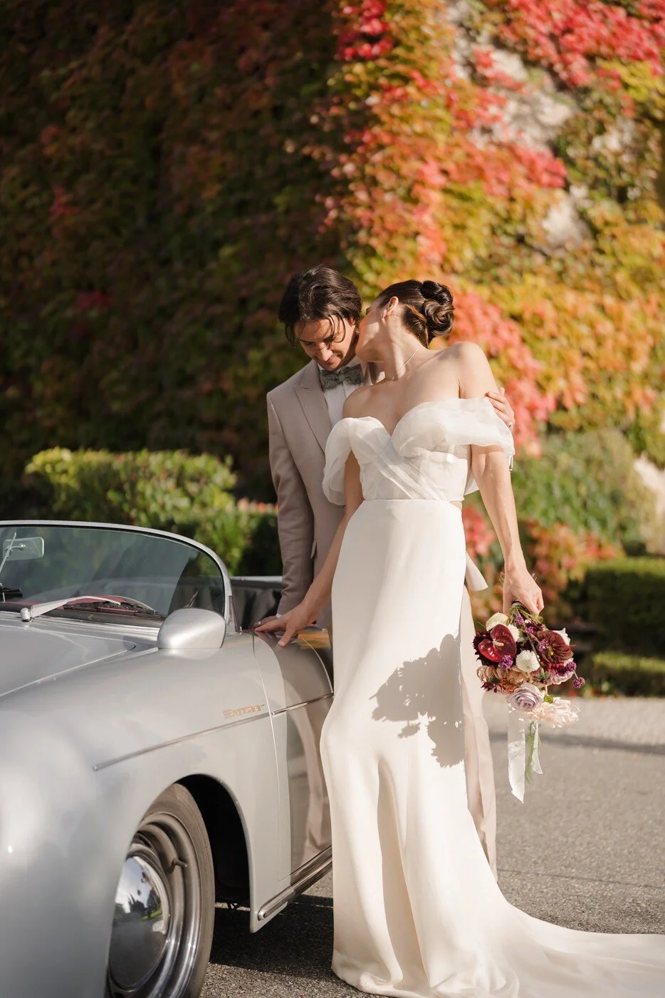 Un couple de mariés partage un moment romantique devant une voiture de collection, avec un bouquet de fleurs en main et un mur de feuillage automnal en arriere-plan