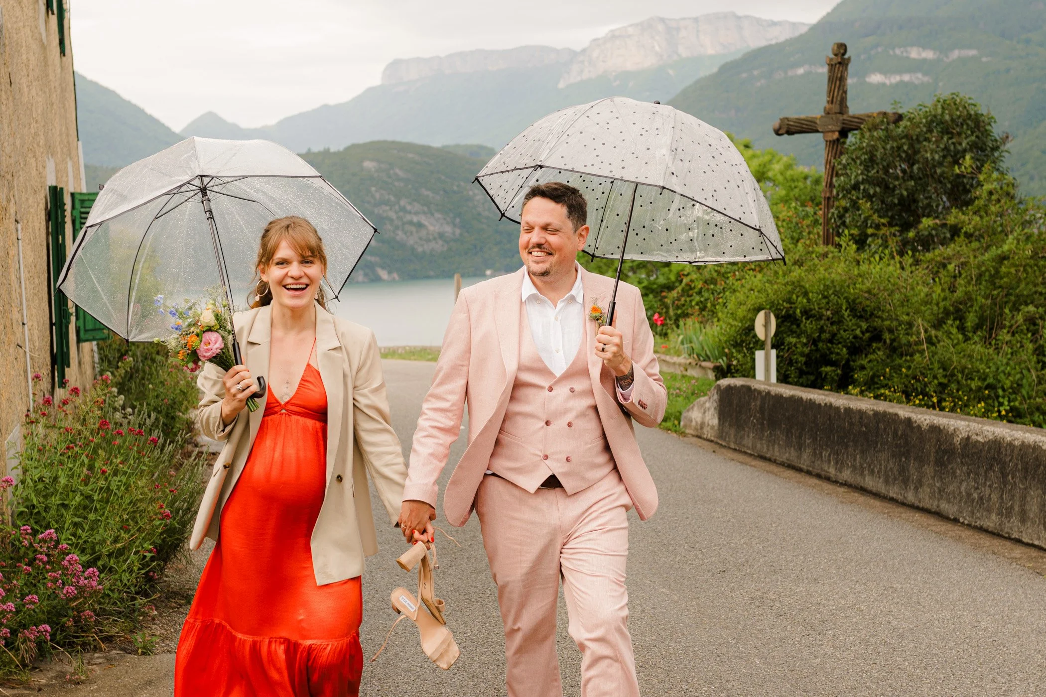 Couple de mariés marchant, main dans la main et tenant des parapluies. Lac et montagne en arrière plan.