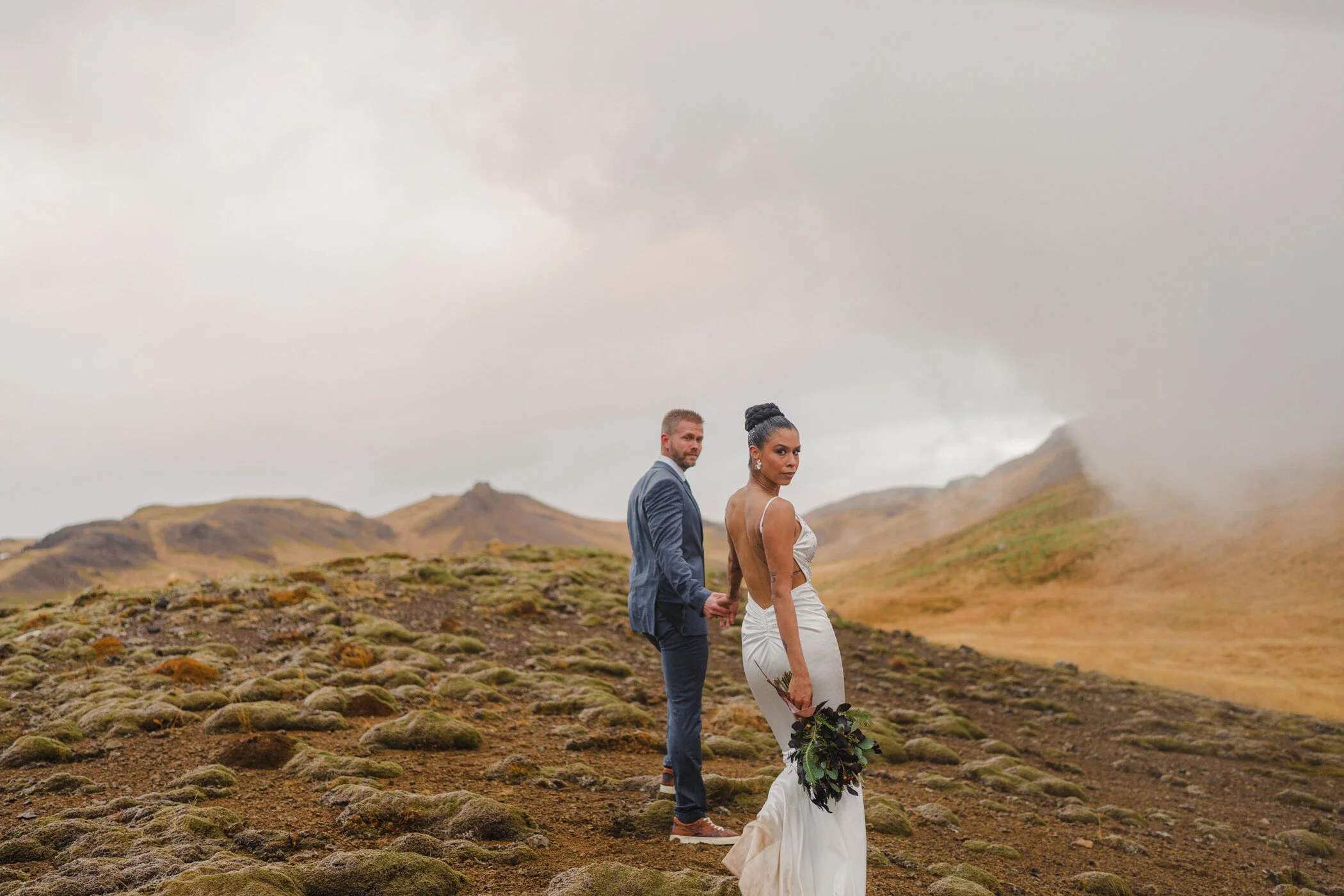 Un couple en tenue de mariage dans un paysage islandais, entouré de montagnes et de nuages, symbolisant l'aventure et l'amour.