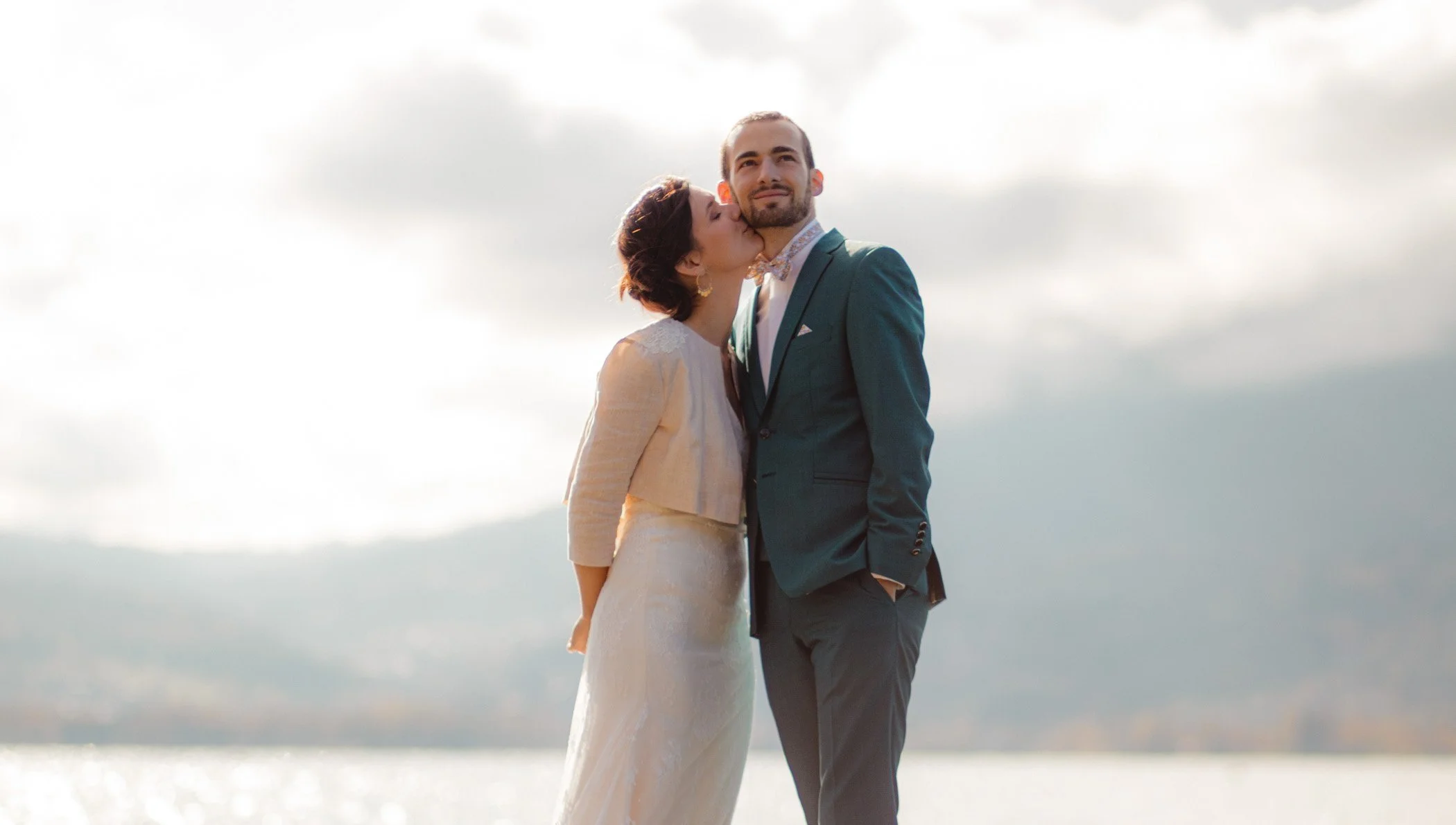 Couple de mariés partage un moment tendre au bord du lac d'Annecy enveloppé par une lumière doucearchant, main dans la main et tenant des parapluies. Lac et montagne en arrière plan.