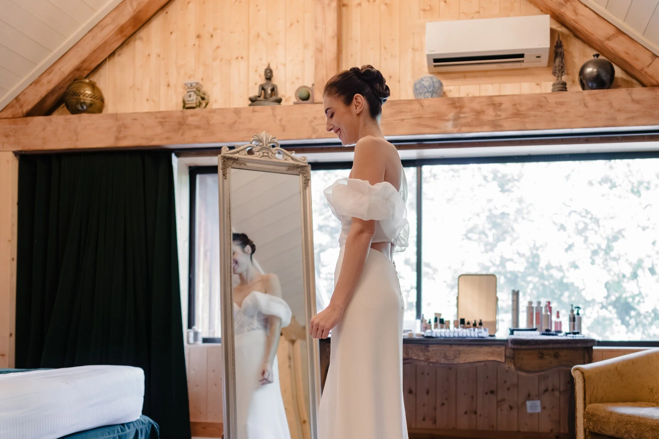 Une mariée, sourire aux lèvres, admire son reflet dans un miroir dans une chambre baignée de lumière naturelle, symbole d'authenticité et d'instantanéité.