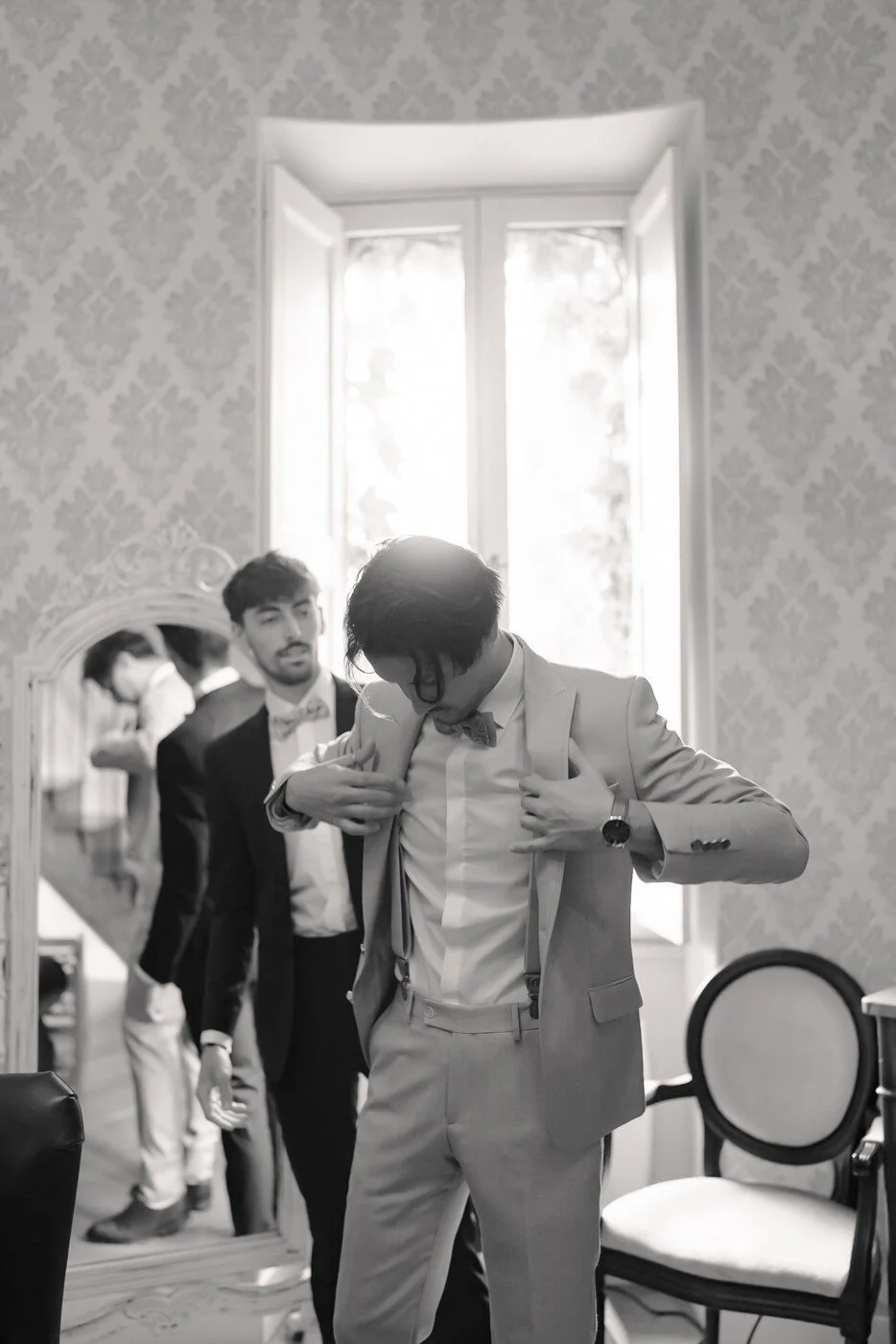 le marié ajuste ses bretelles avec l'aide de ses témoins, baigné dans une lumière douce, avant la cérémonie.