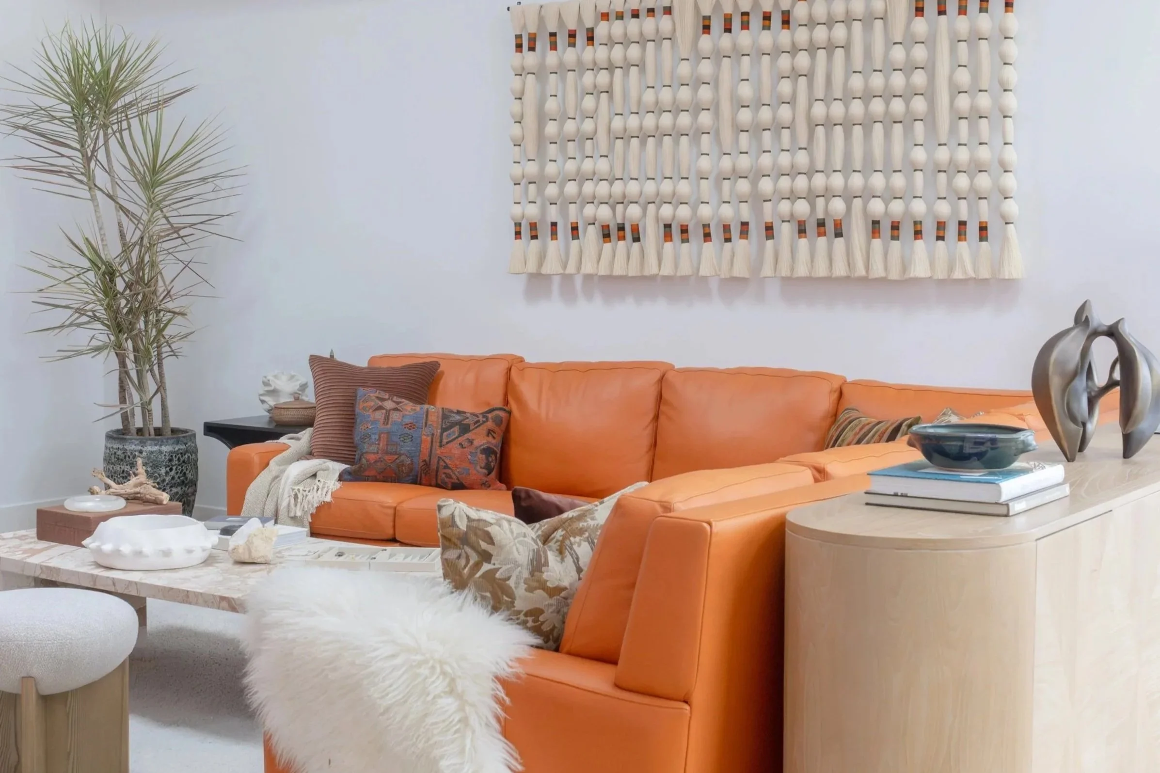 Living room with orange leather sofa, decorative pillows, a white fluffy throw, a marble coffee table with sculptures and shells, a potted plant, and a wall hanging with wooden beads and tassels.