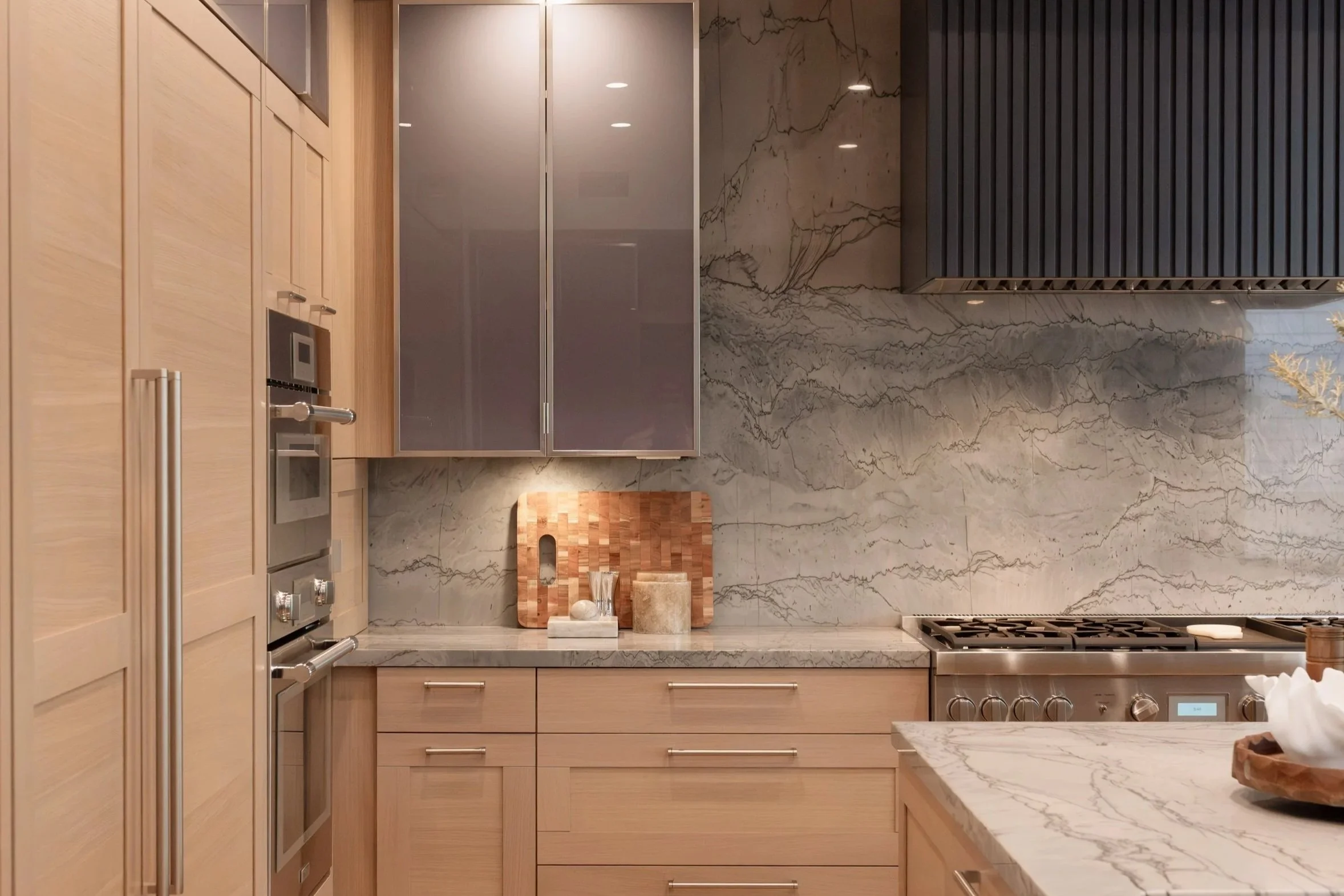 Modern kitchen with light wood cabinets, marble countertops, and stainless steel appliances, with wood flooring and a black vent hood.