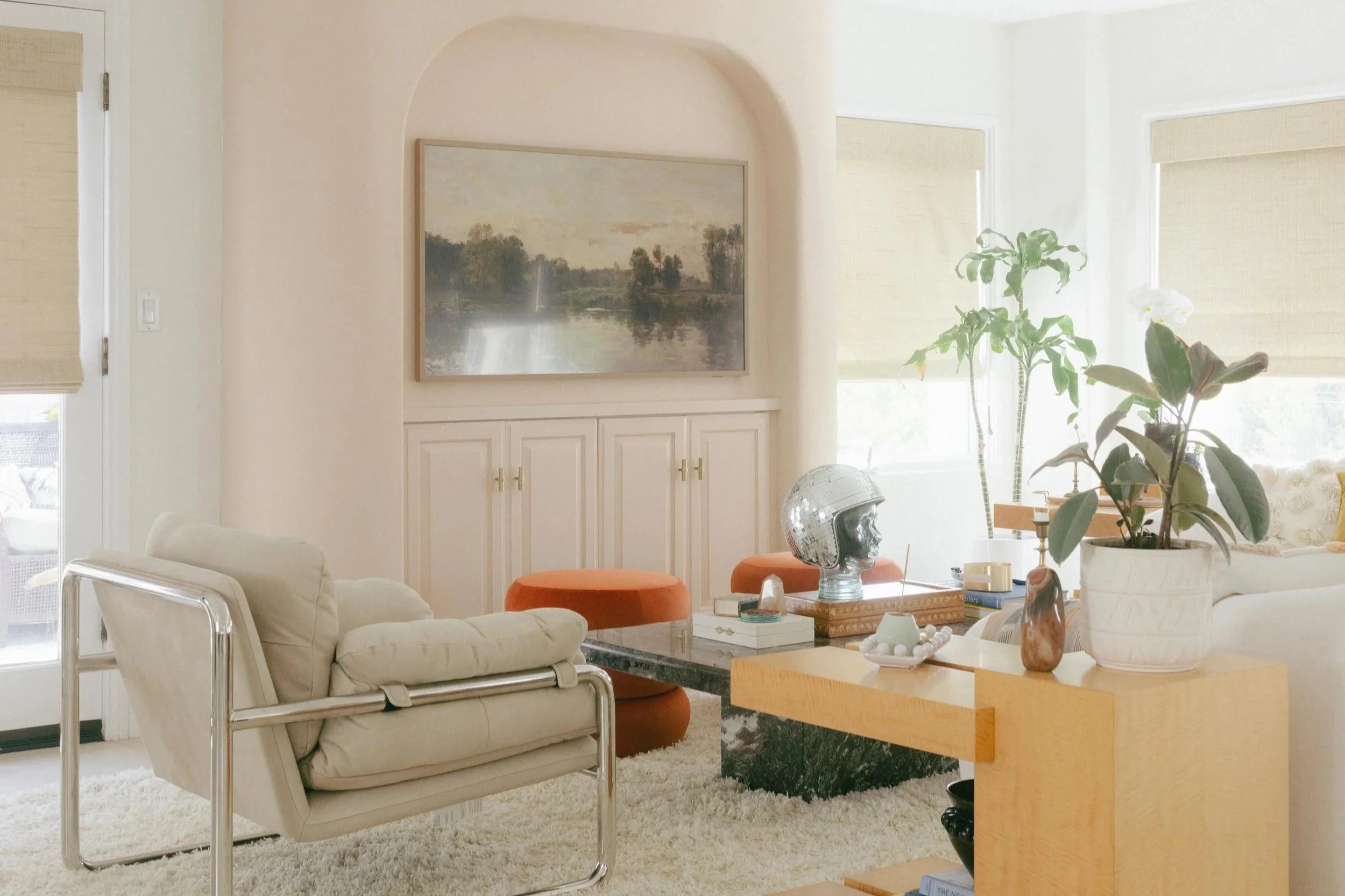 Bright living room with white walls and large windows with beige blinds. Central area has a white armchair with chrome frame, a cream-colored couch, and a black coffee table. Decor includes a silver helmet, books, and a large potted plant. A painting of a riverside landscape hangs on the wall.