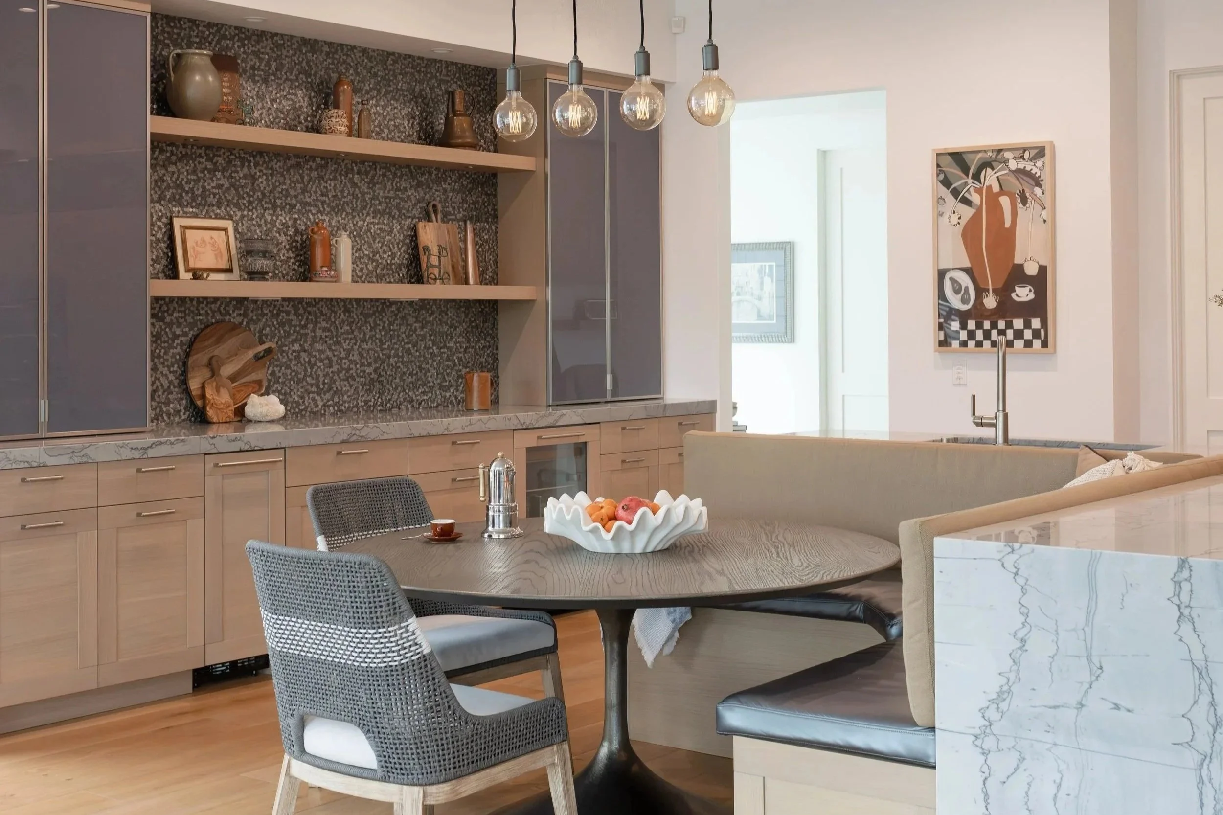 Modern kitchen and dining area with a round table, chairs, a built-in bench, and a marble counter, featuring decorative items and hanging light bulbs.