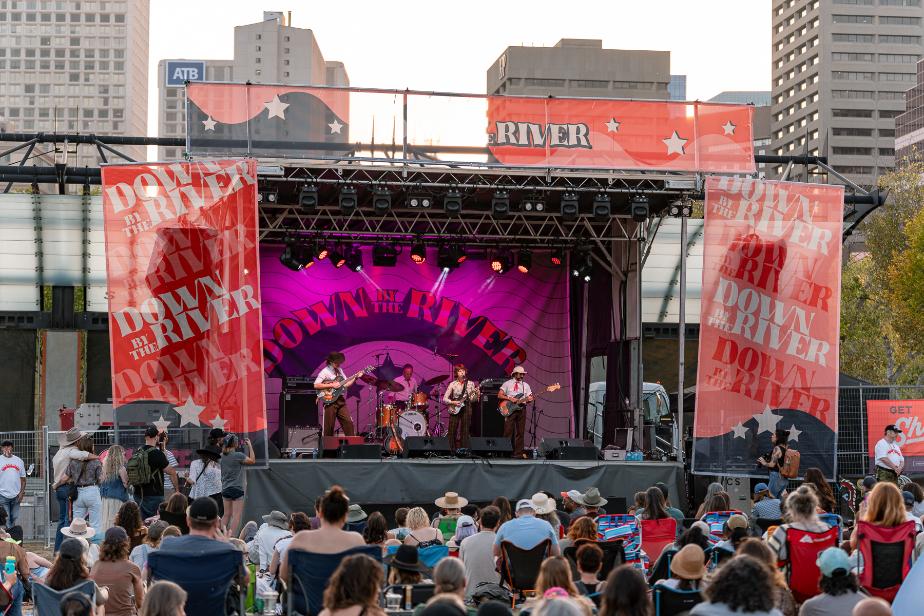 A live outdoor concert on a stage with musicians performing and an audience seated in chairs and on the ground, with city buildings in the background.
