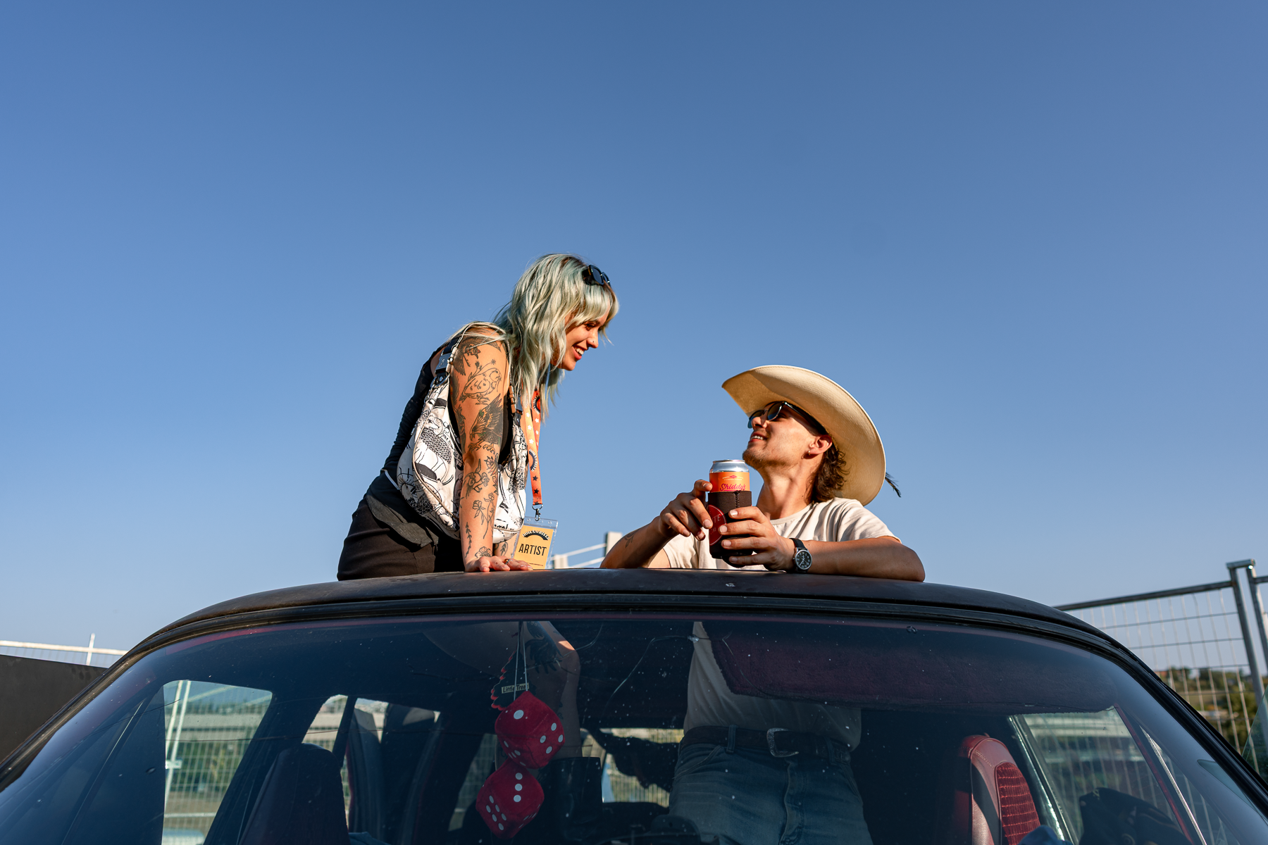 Two people, a woman and a man, are enjoying a drink from a soda can while standing and leaning on the roof of a car during daytime with a clear blue sky.