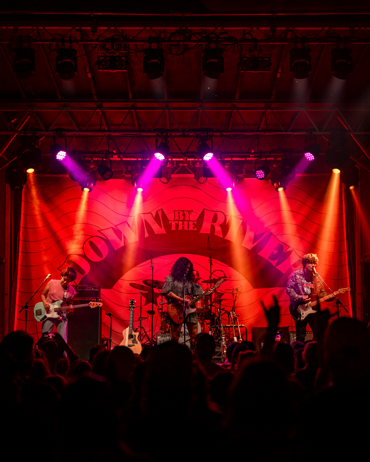 A band performing on stage with pink and yellow stage lighting, in front of a large red backdrop with text that reads 'DOWN BY THE RIVER.' The audience is visible in the foreground.