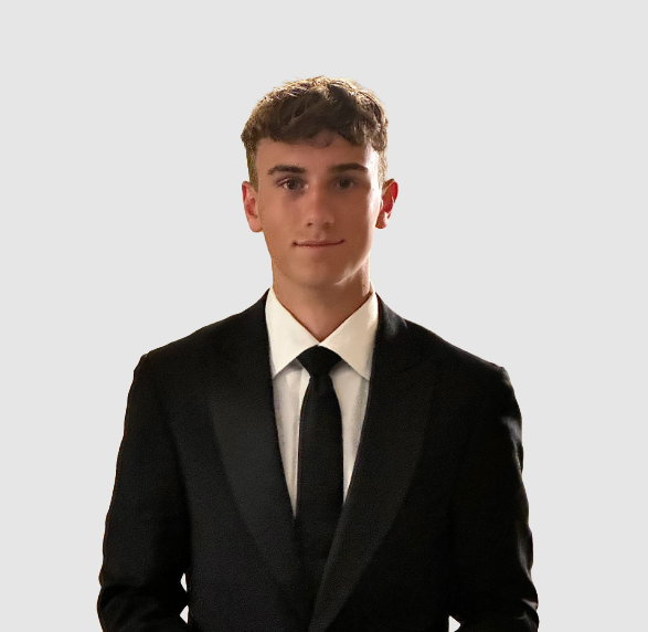 Young man in black suit, white shirt, and black tie standing against a plain light background.