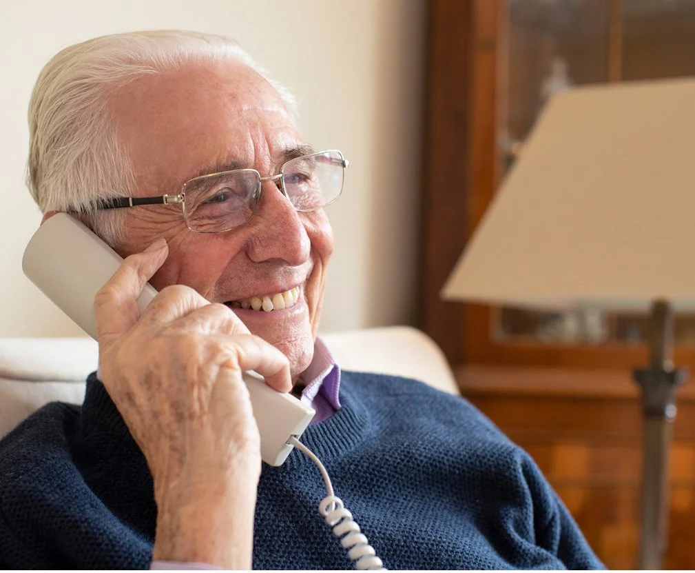 An elderly man with gray hair, wearing glasses and a navy sweater, is smiling while talking on a landline phone in a cozy indoor setting.