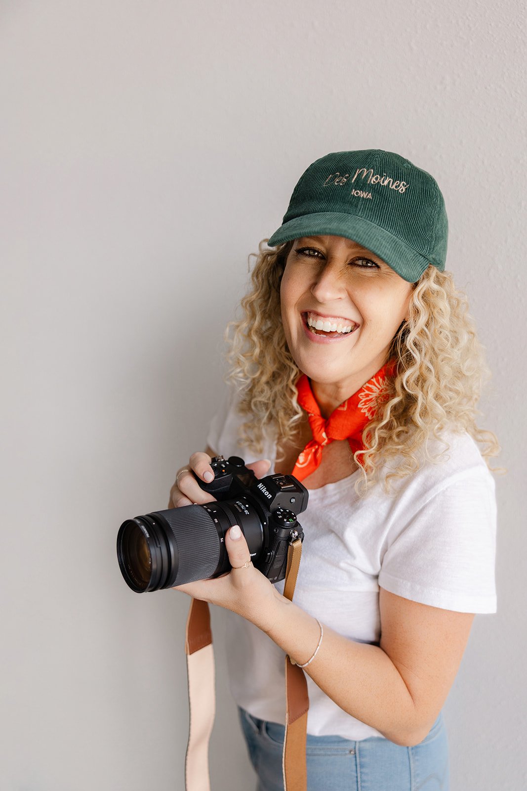 Jill wearing a red bandana and dark green baseball cap embroidered with Des Moines Iowa and holding her camera smiling