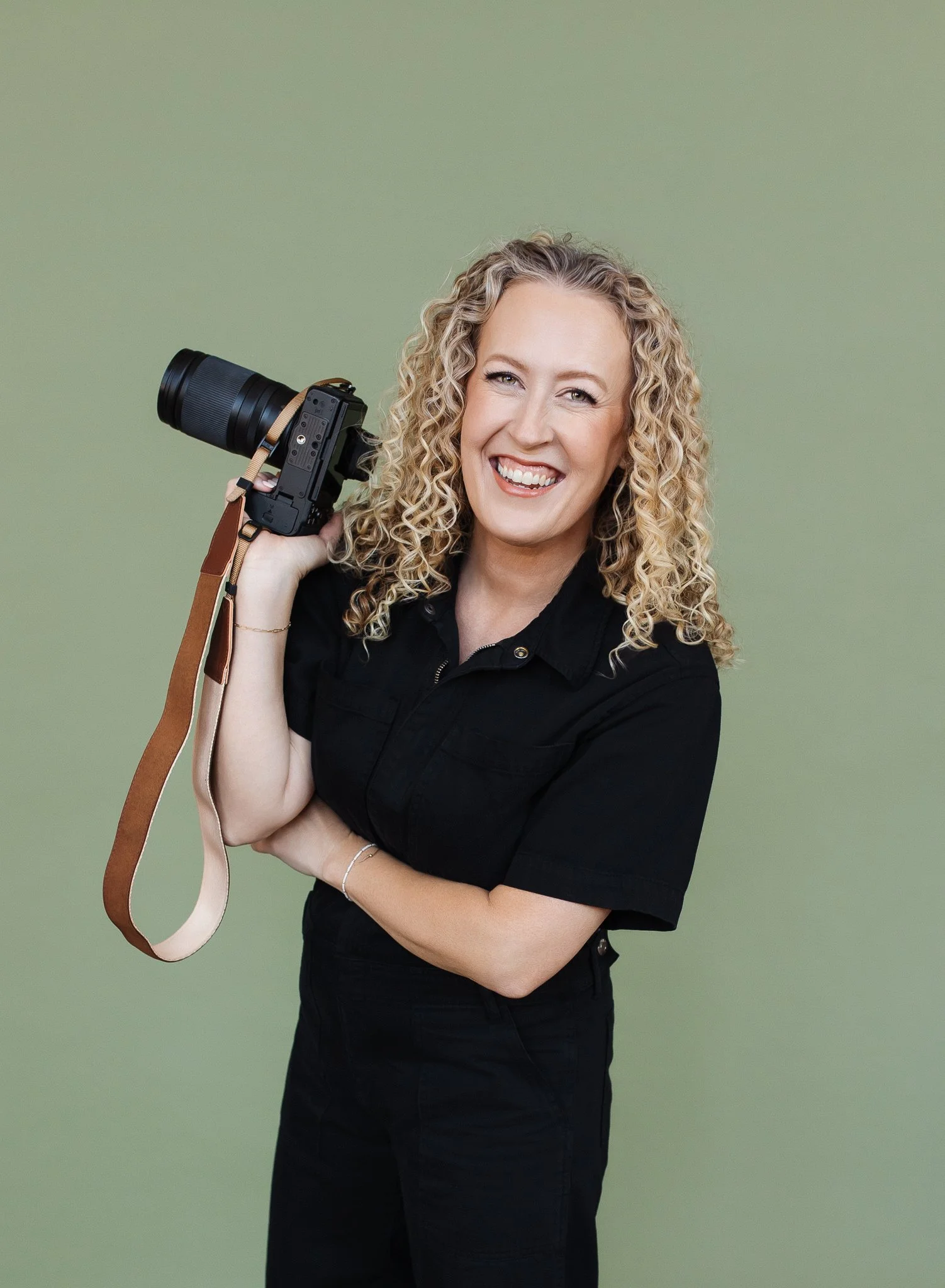 Jill Neil with curly blond hair holding up her camera in one hand while smiling for a lifestyle headshot in front of a green studio backdrop