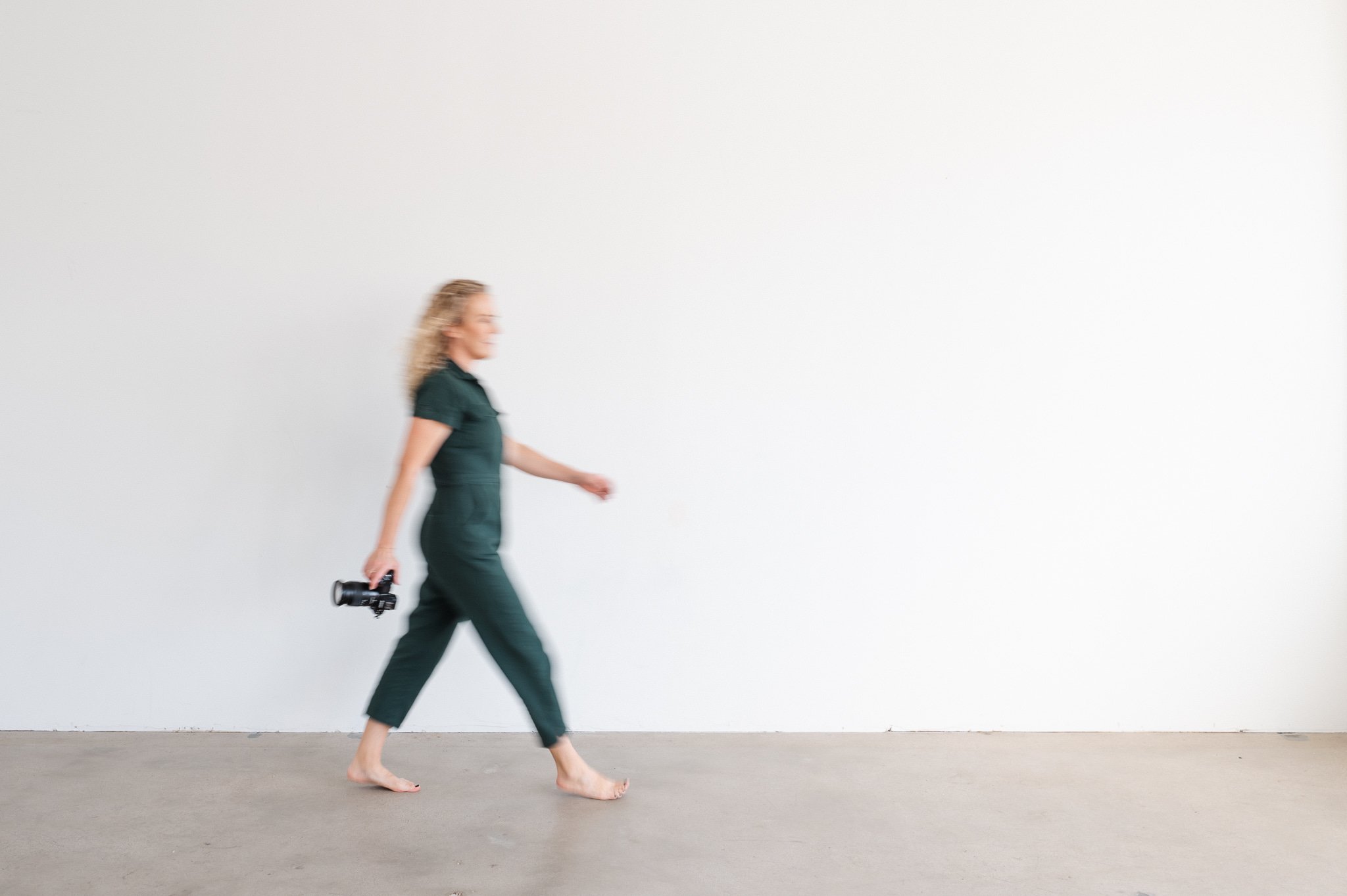 Jill Neil with camera in hand walking across concrete studio floor in front of white wall