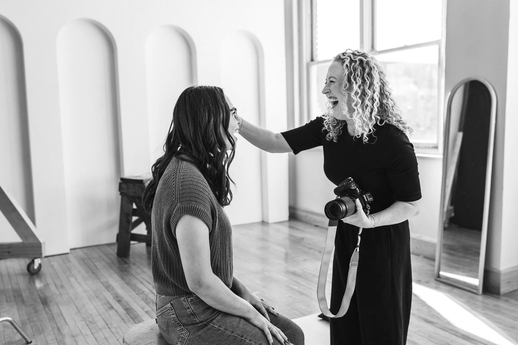Jill Neil laughing in photo studio while adjusting her client's hair with one hand and holding her camera with the other