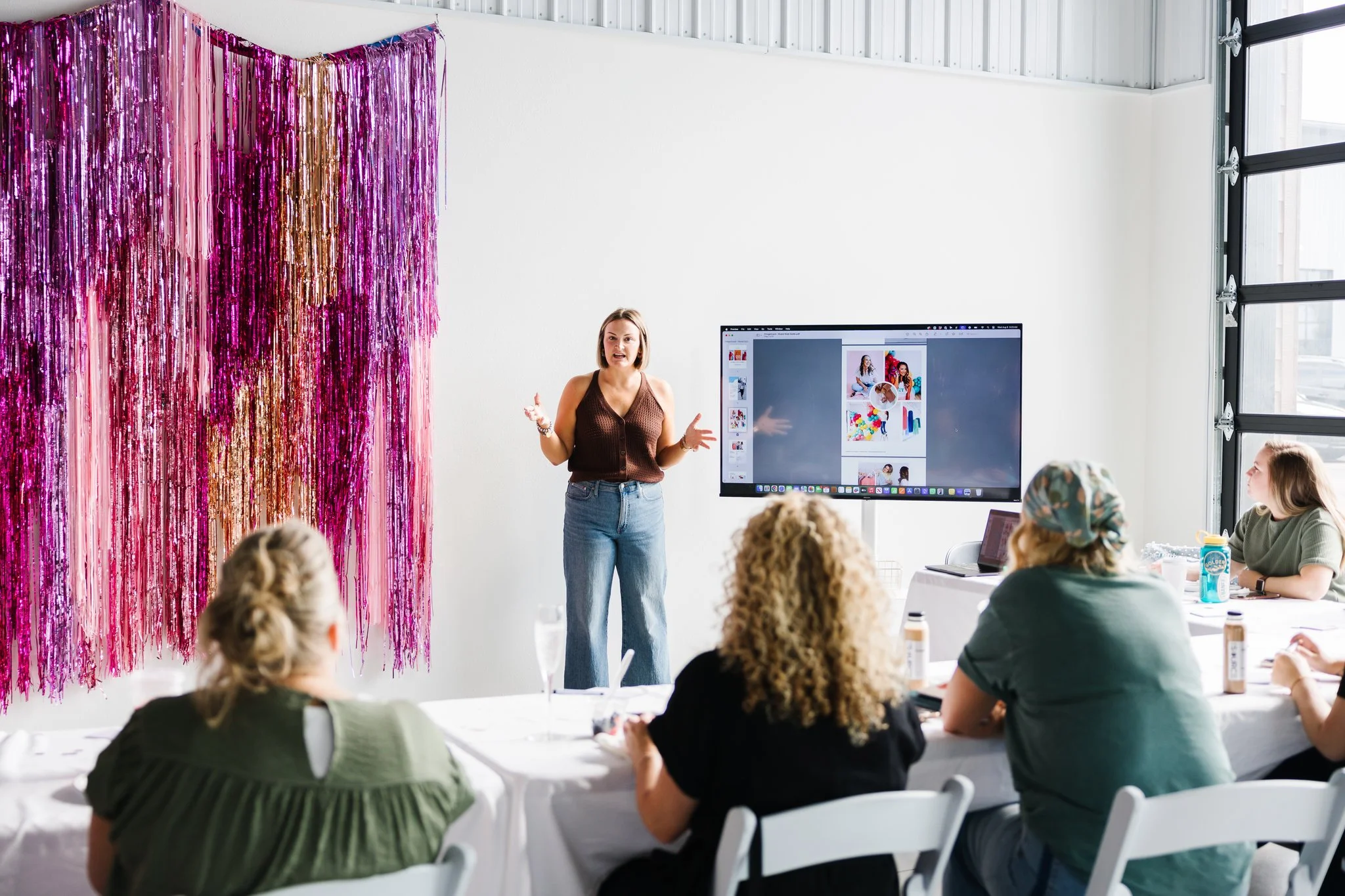 Maddie Peschong standing between a shiny tinsel backdrop and a large tv monitor teaching a table of females in her photography studio