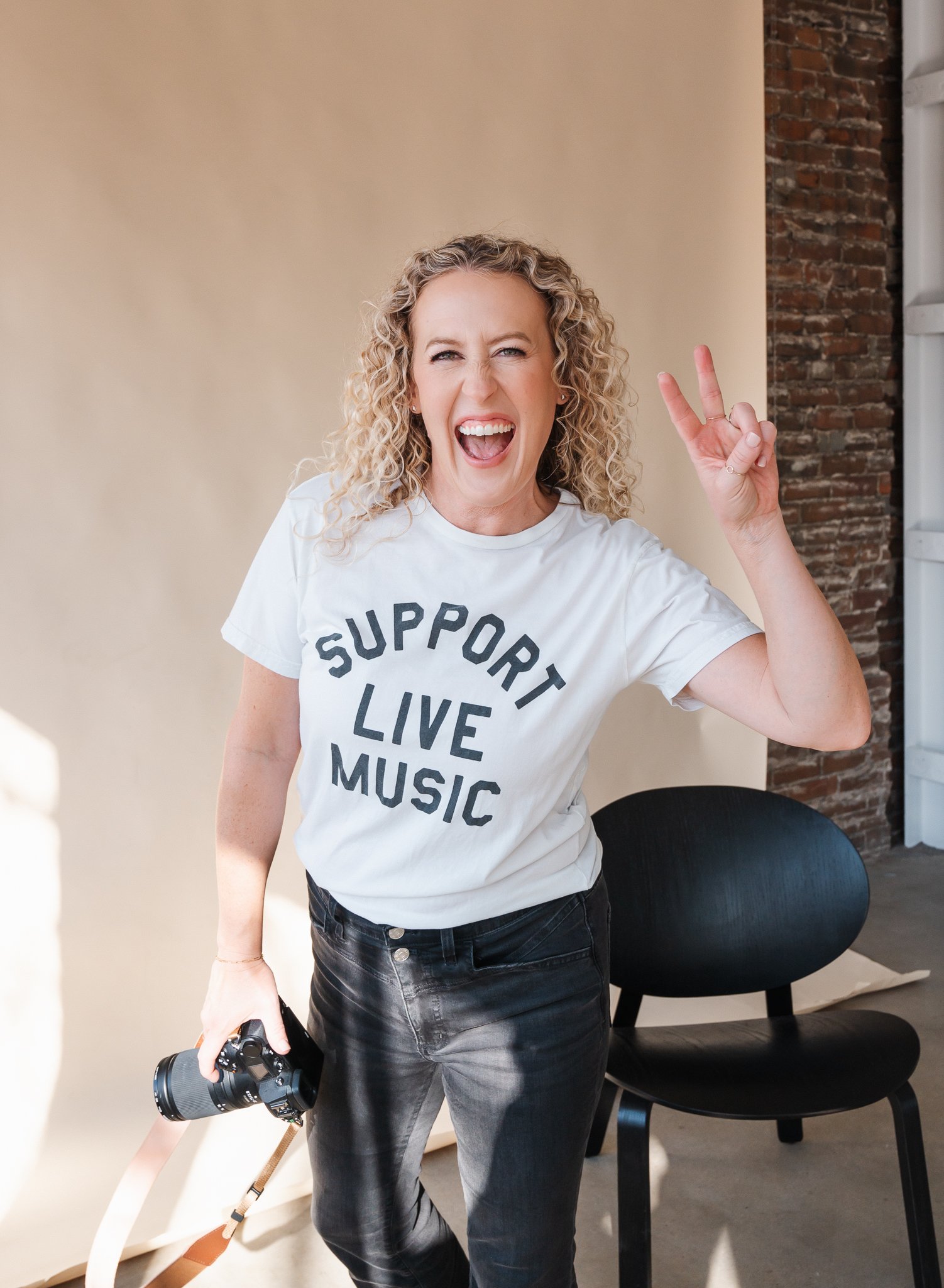 Jill wearing a t-shirt that says Support Live Music while holding camera in one hand and flashing a peace sign with other and laughing
