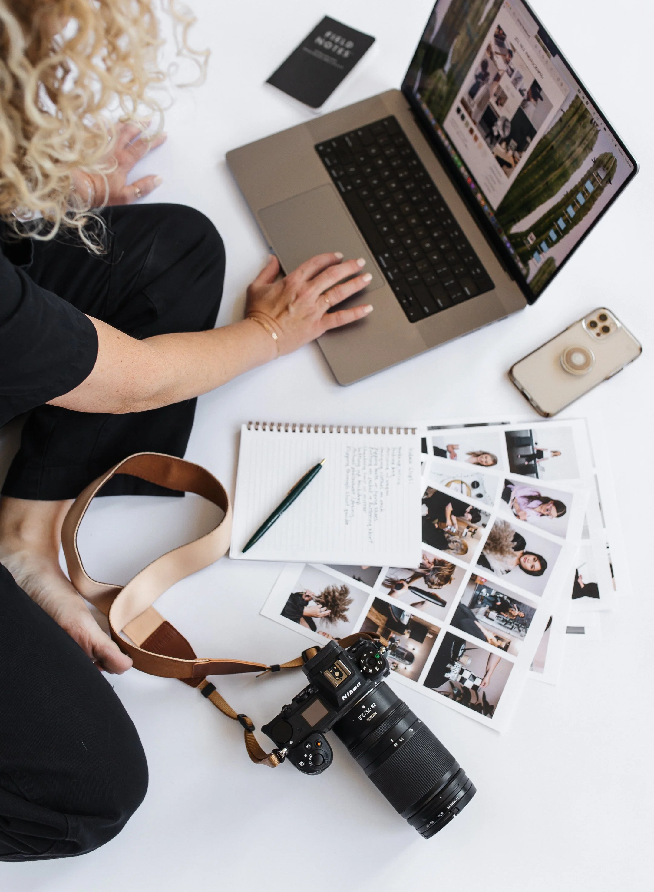 Jill sitting on floor reaching for laptop surrounded by her camera, notebook, phone and photo prints