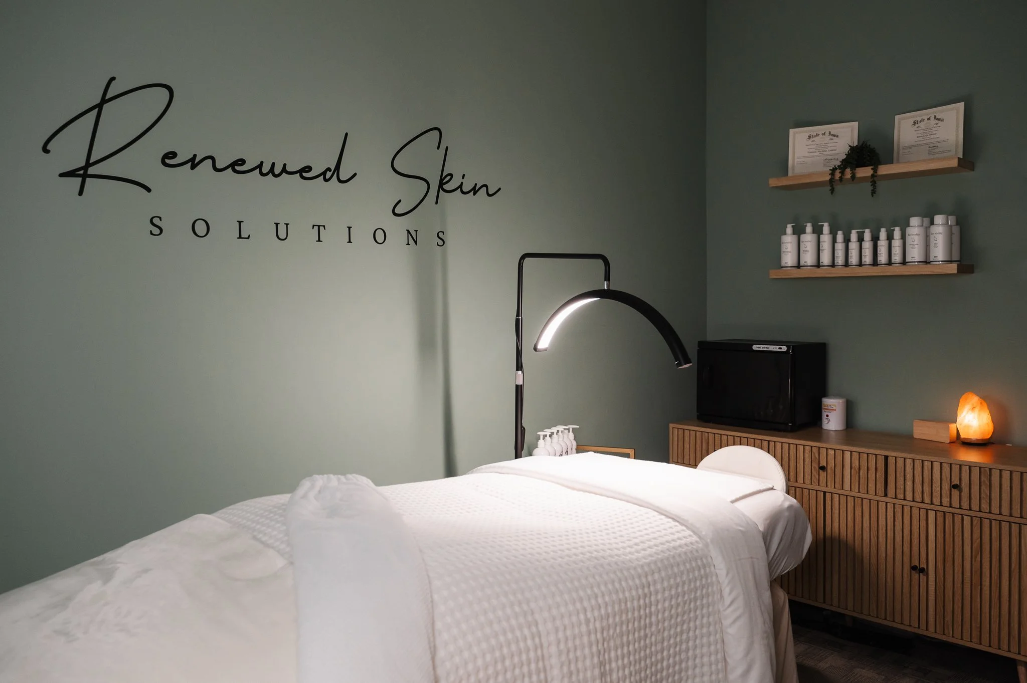 facial treatment bed in soothing dark room with glow of lamps and Renewed Skin Solutions logo on sage green wall