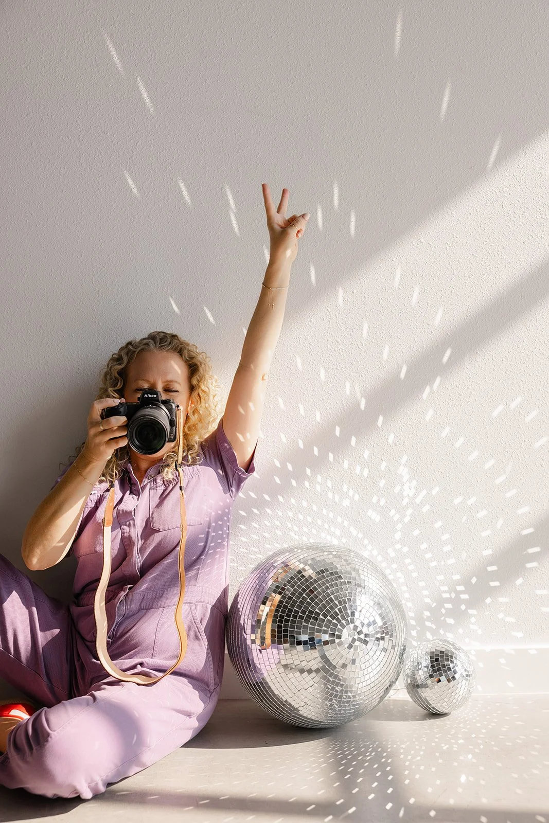 Jill sitting on floor next to large disco balls reflecting onto wall holding her camera in one hand and flashing a peace sign with the other