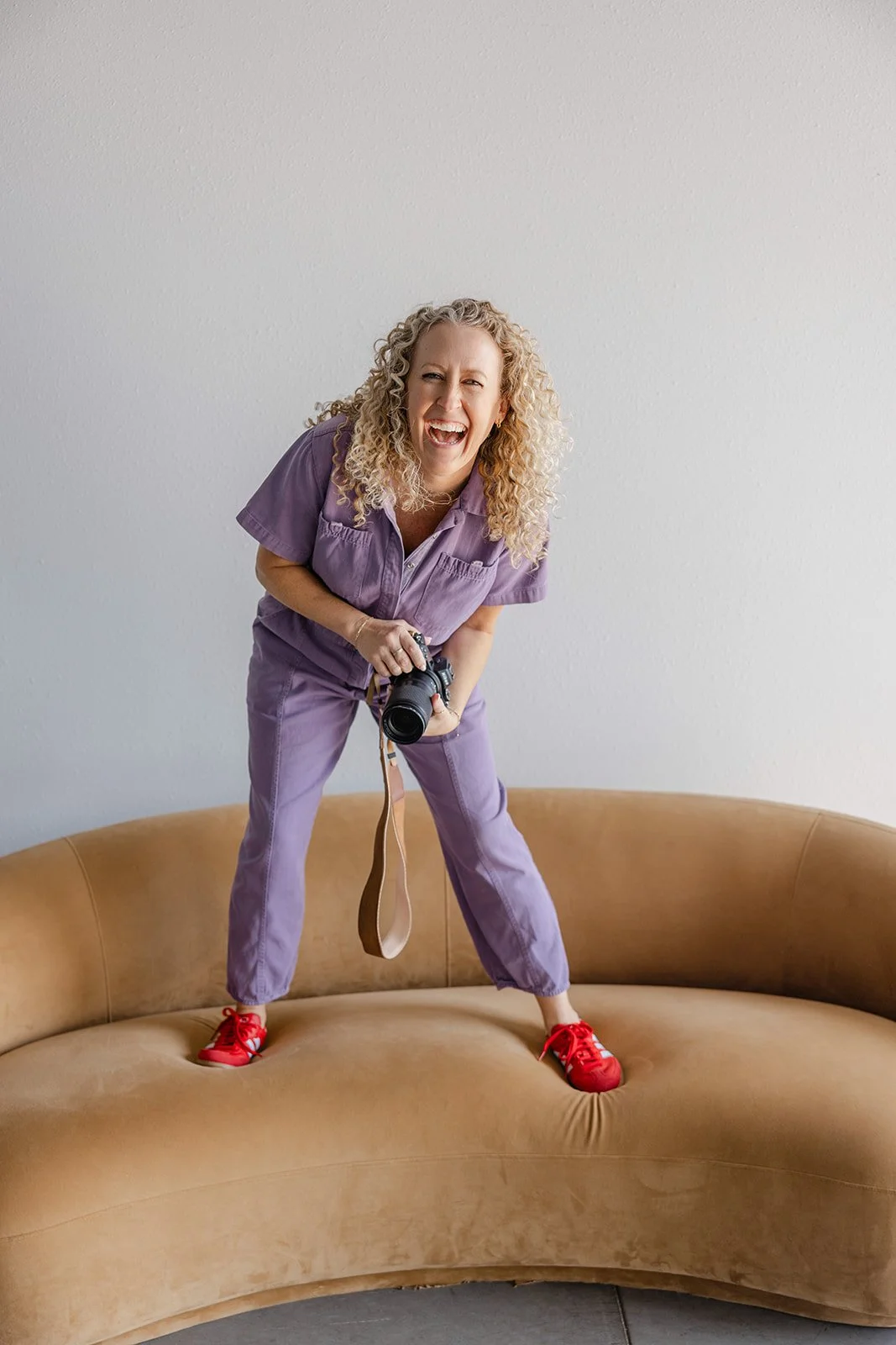 Jill standing on couch in a purple jumpsuit holding camera bending over laughing with her curly hair falling over her shoulders