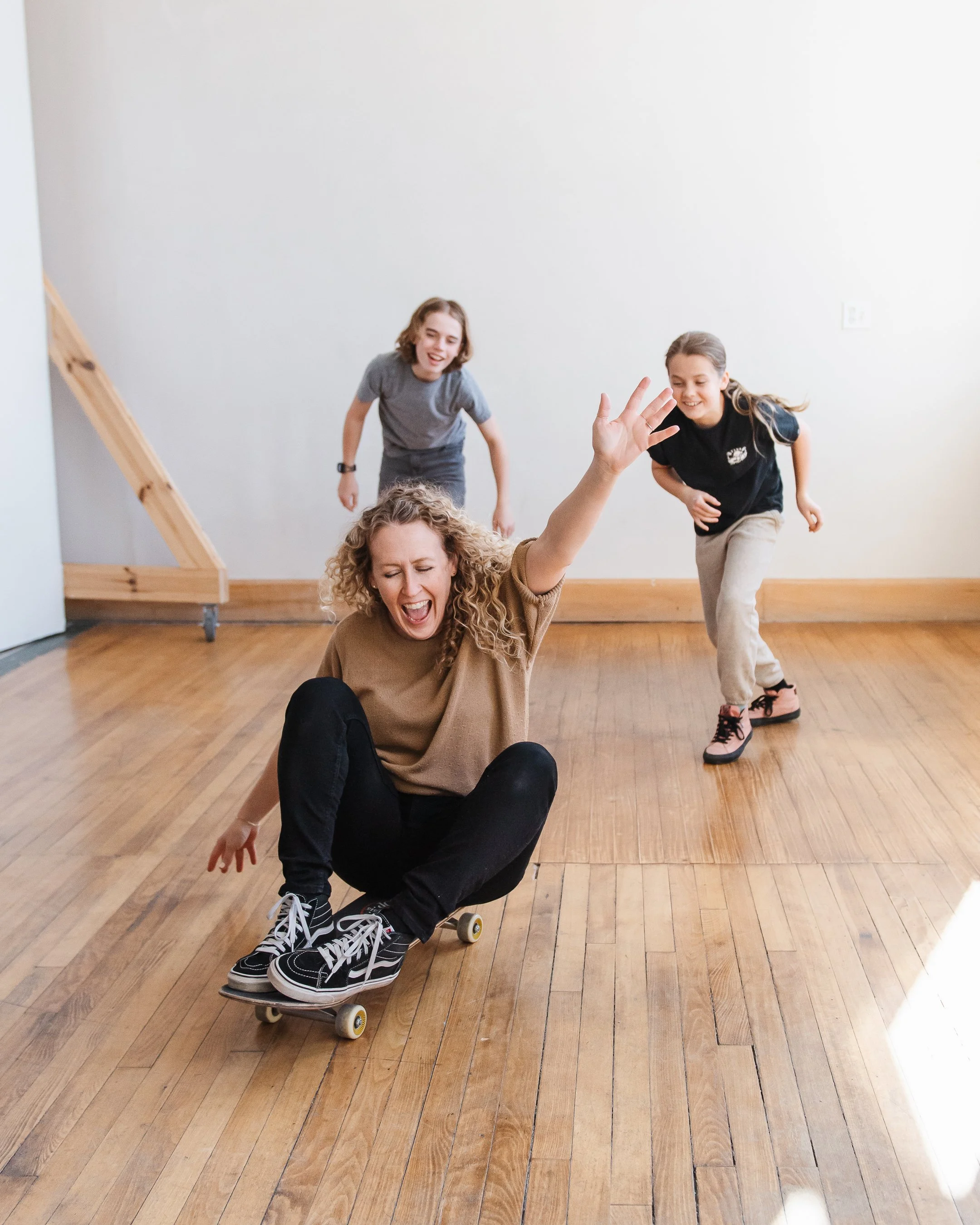 Jill seated on a skateboard riding across the photo studio floor with arm up in air and her two kids chasing her