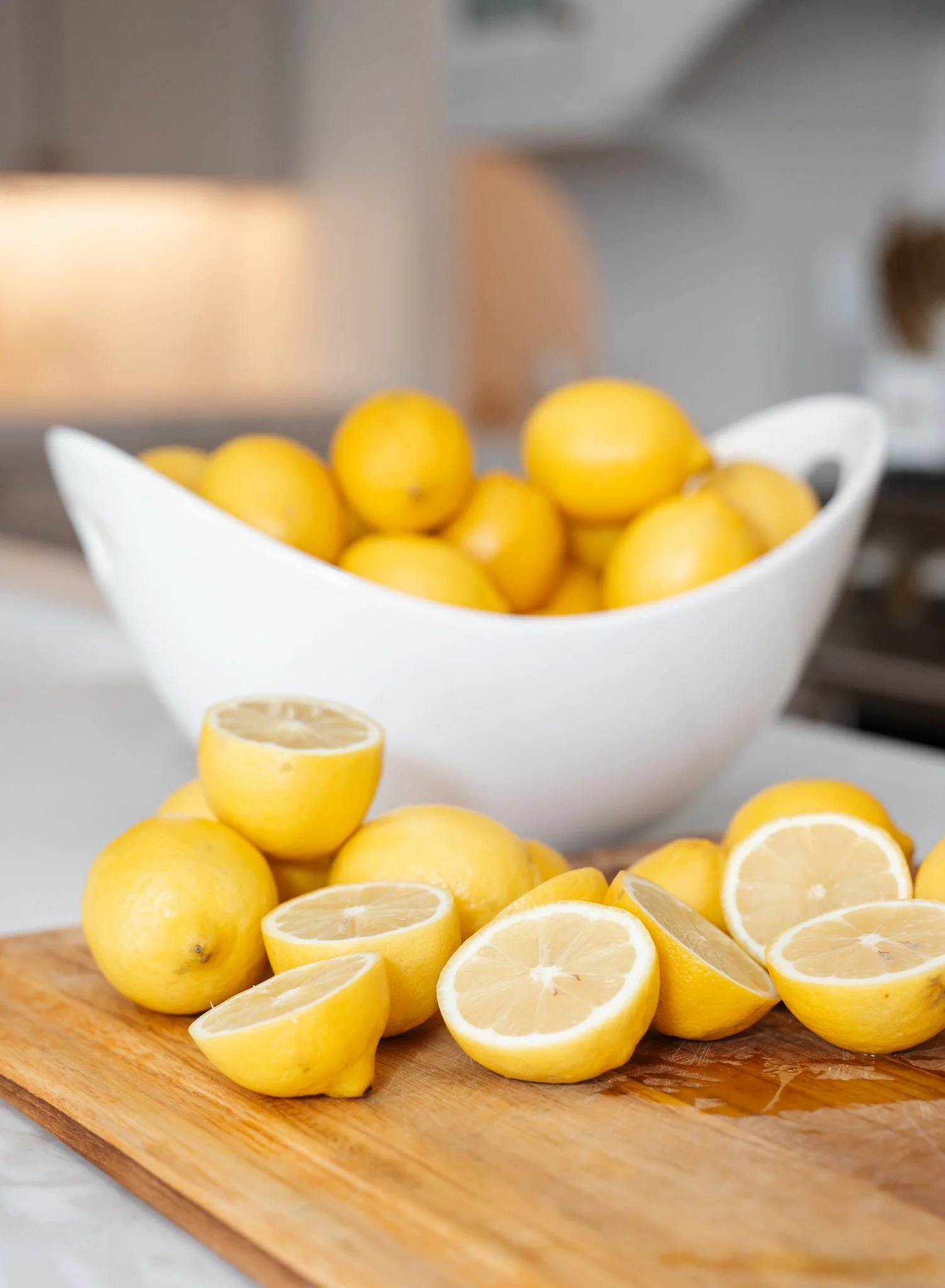 multiple lemons cut in half on cutting board with large bowl of whole lemons on counter behind it