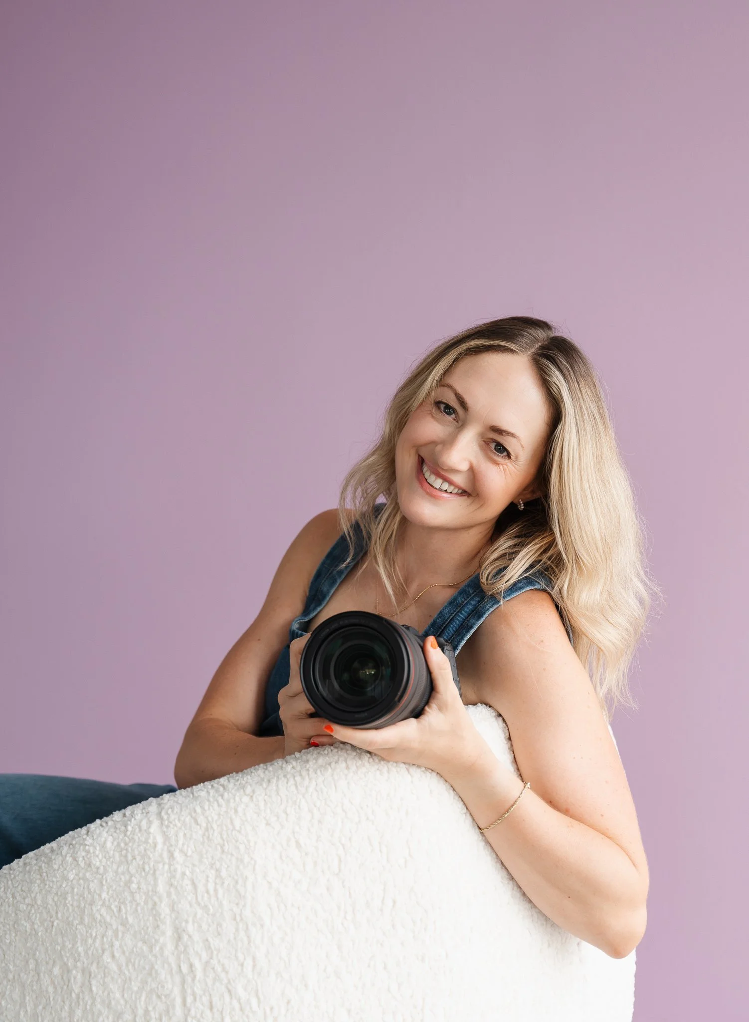 female photographer sitting with her arm over the back of a white chair holding her camera in front of a lilac purple studio backdrop