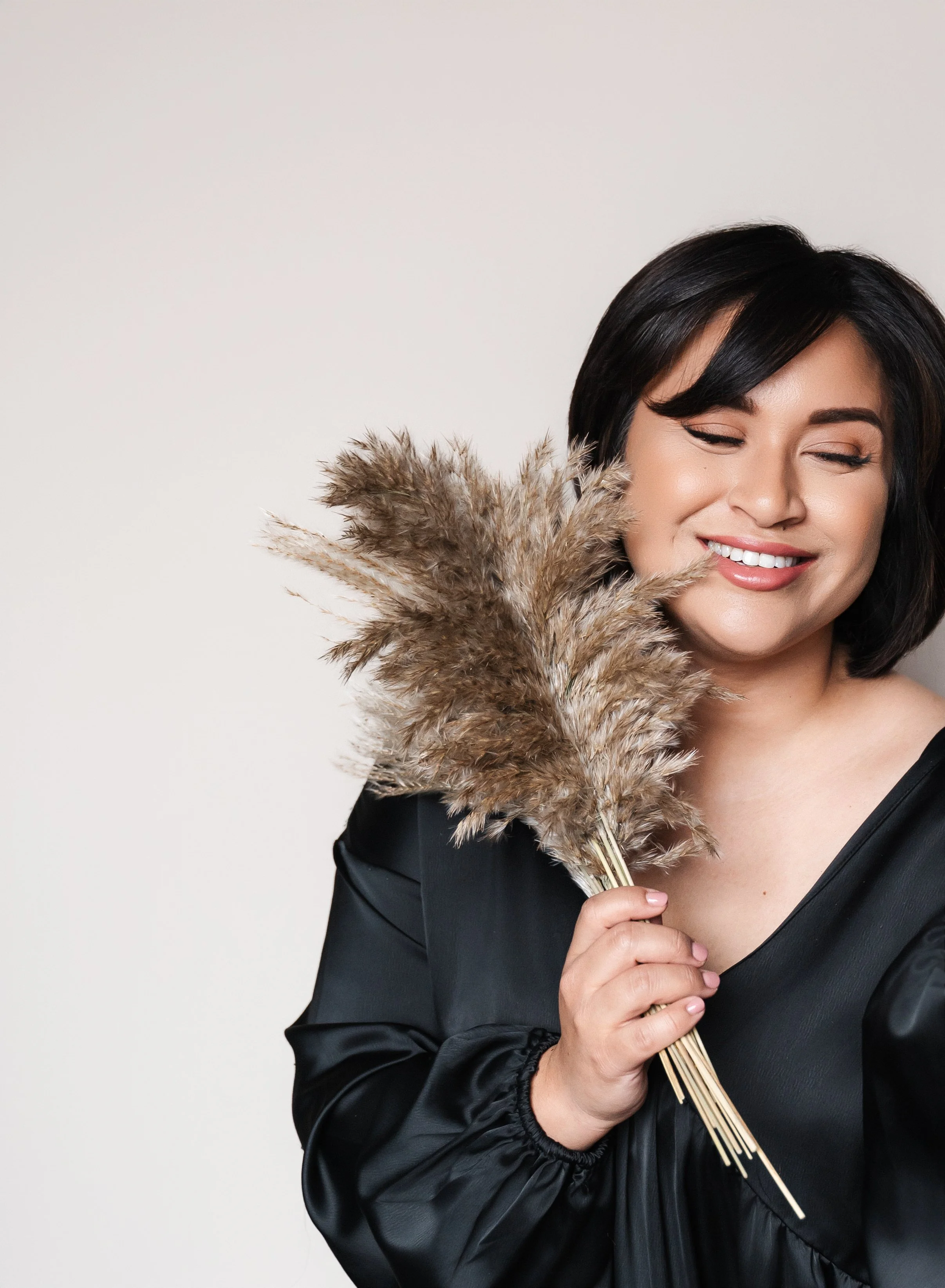 female with short dark hair holding dried florals up to her face smiling with her eyes closed