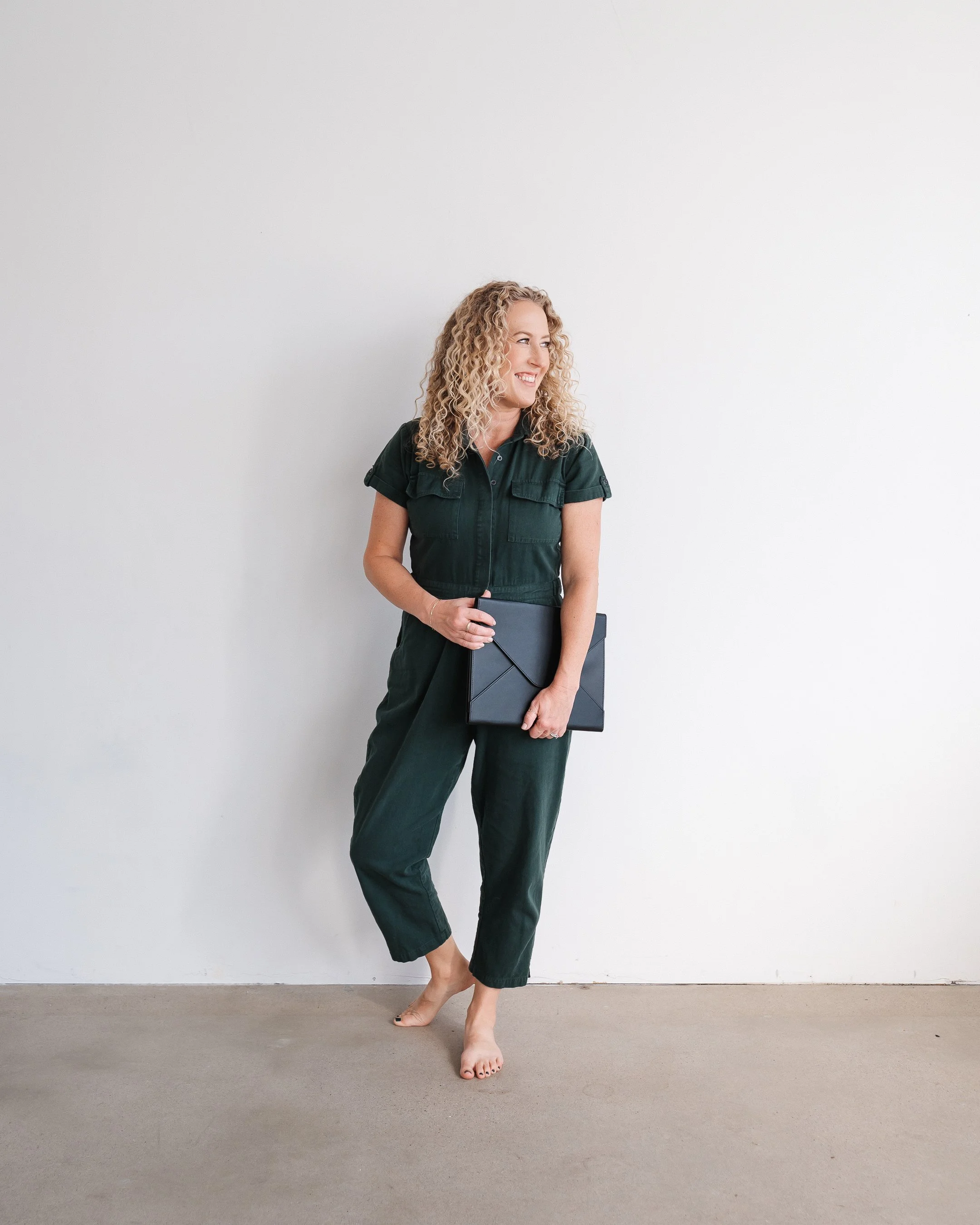 Jill wearing a dark green jumpsuit standing against white studio wall holding portfolio and smiling