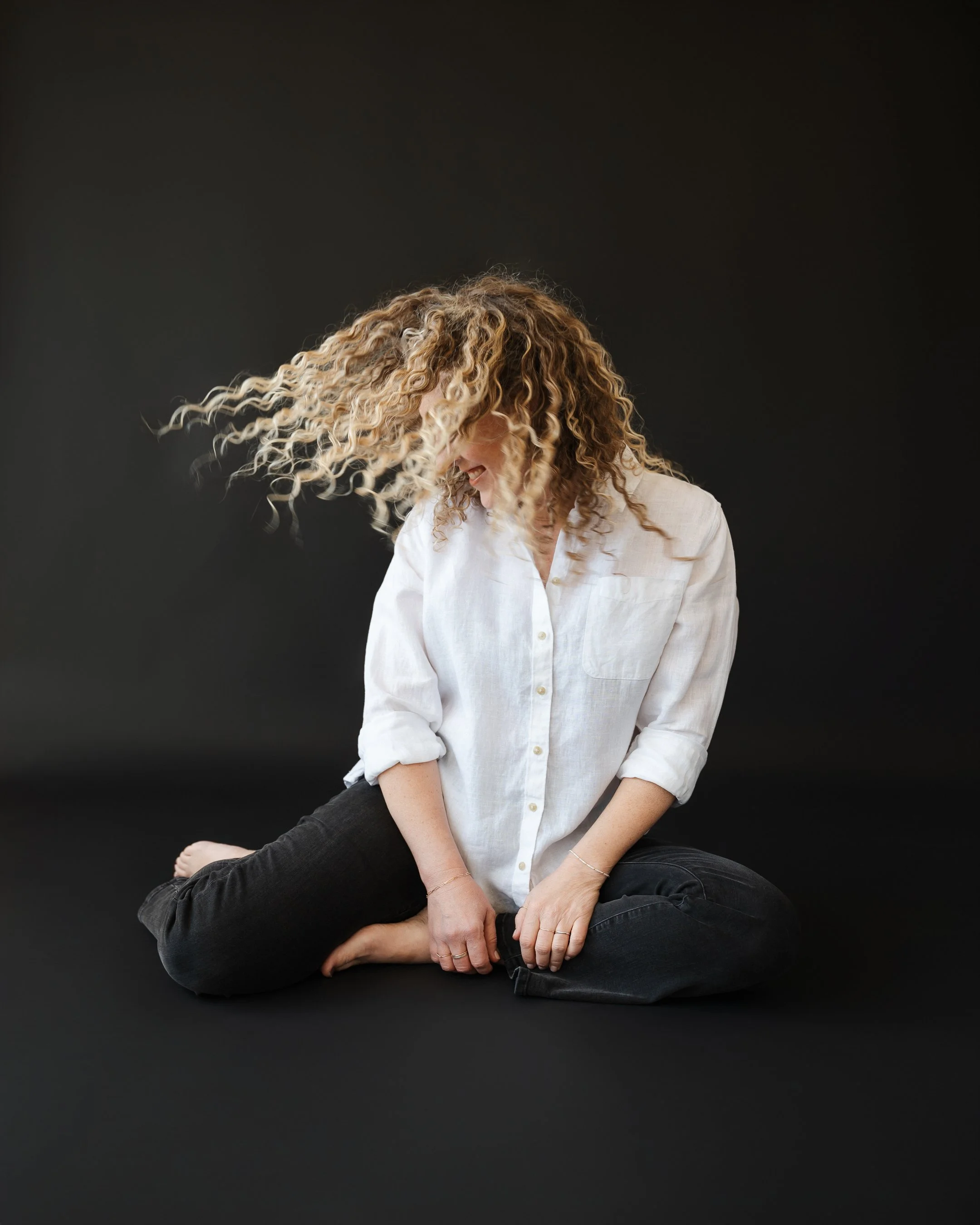 Jill sitting on black seamless backdrop whipping her blond natural curls around her face