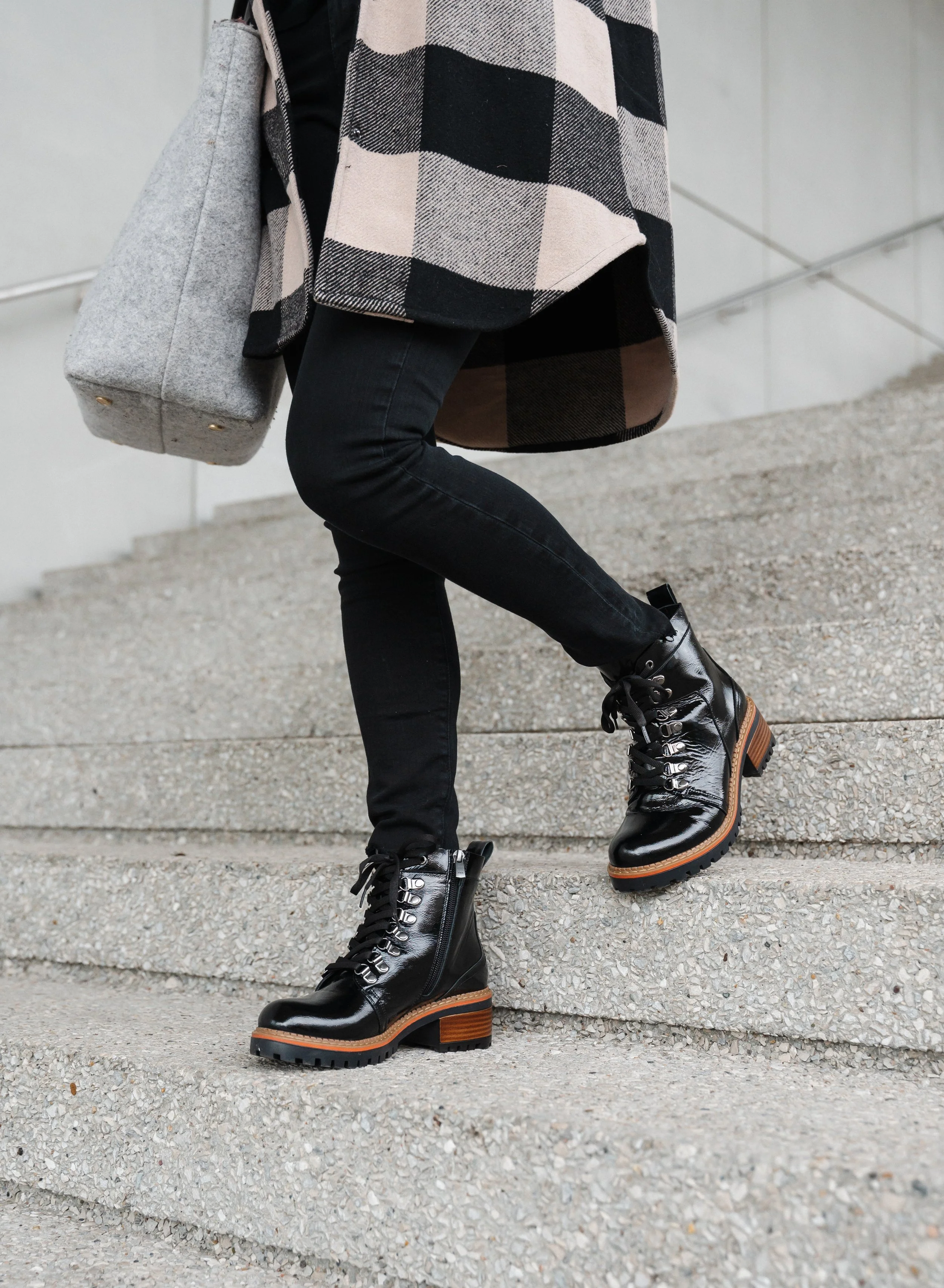cropped half body photo of female walking down stairs in shiny black leather boot with laces wearing black jeans and long flannel checked jacket and tote bag