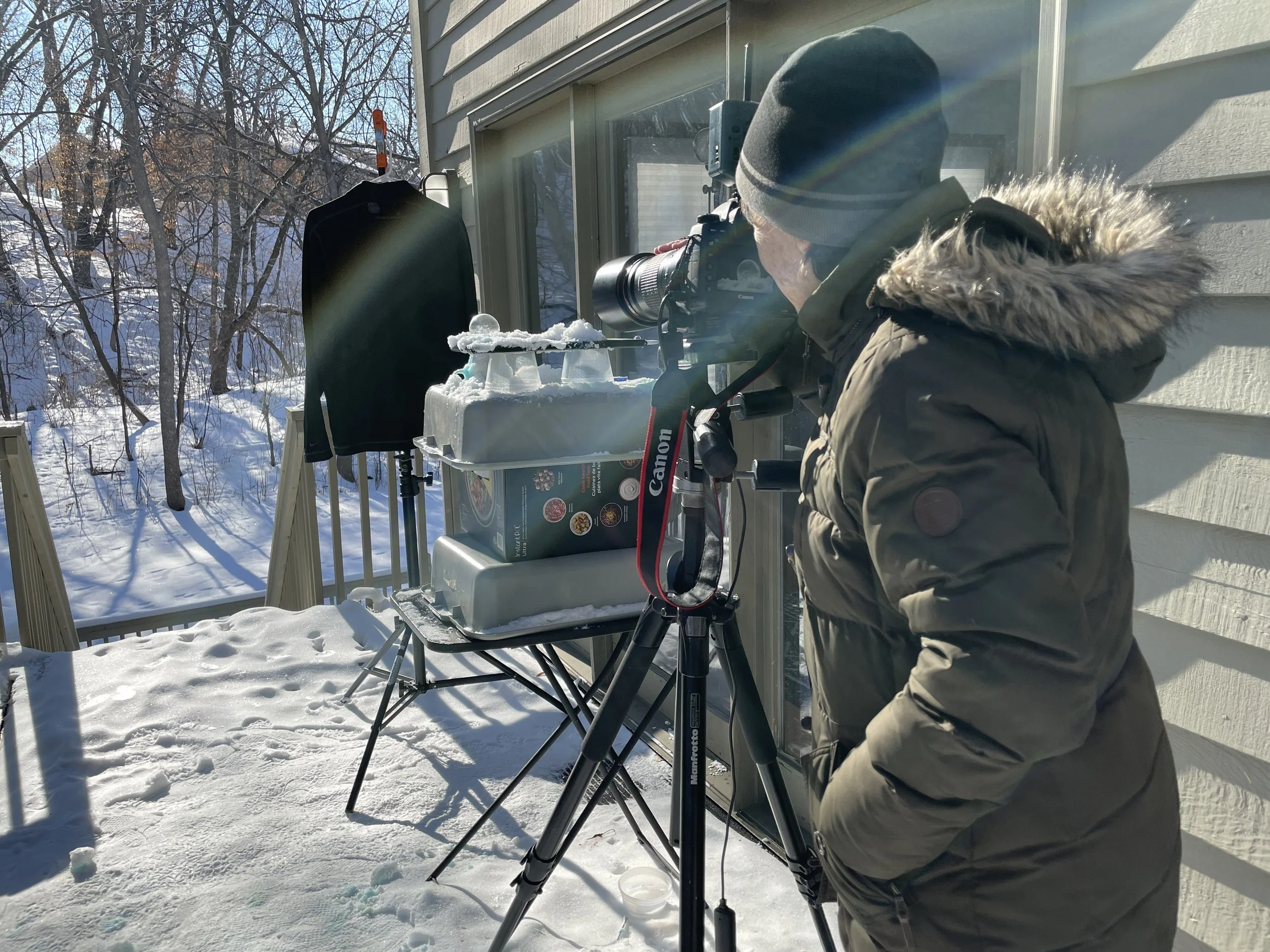 A person in winter clothing, including a beanie and a coat with fur-lined hood, is looking through a camera mounted on a tripod outside on a snowy day. There are snow-covered trees and a house exterior in the background, and sunlight is creating a rainbow flare across the image.
