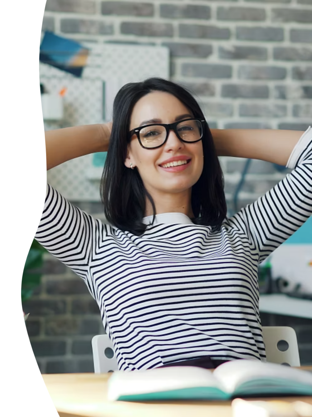 A young woman with black hair and glasses smiling, sitting at a desk in an office with a brick wall background.