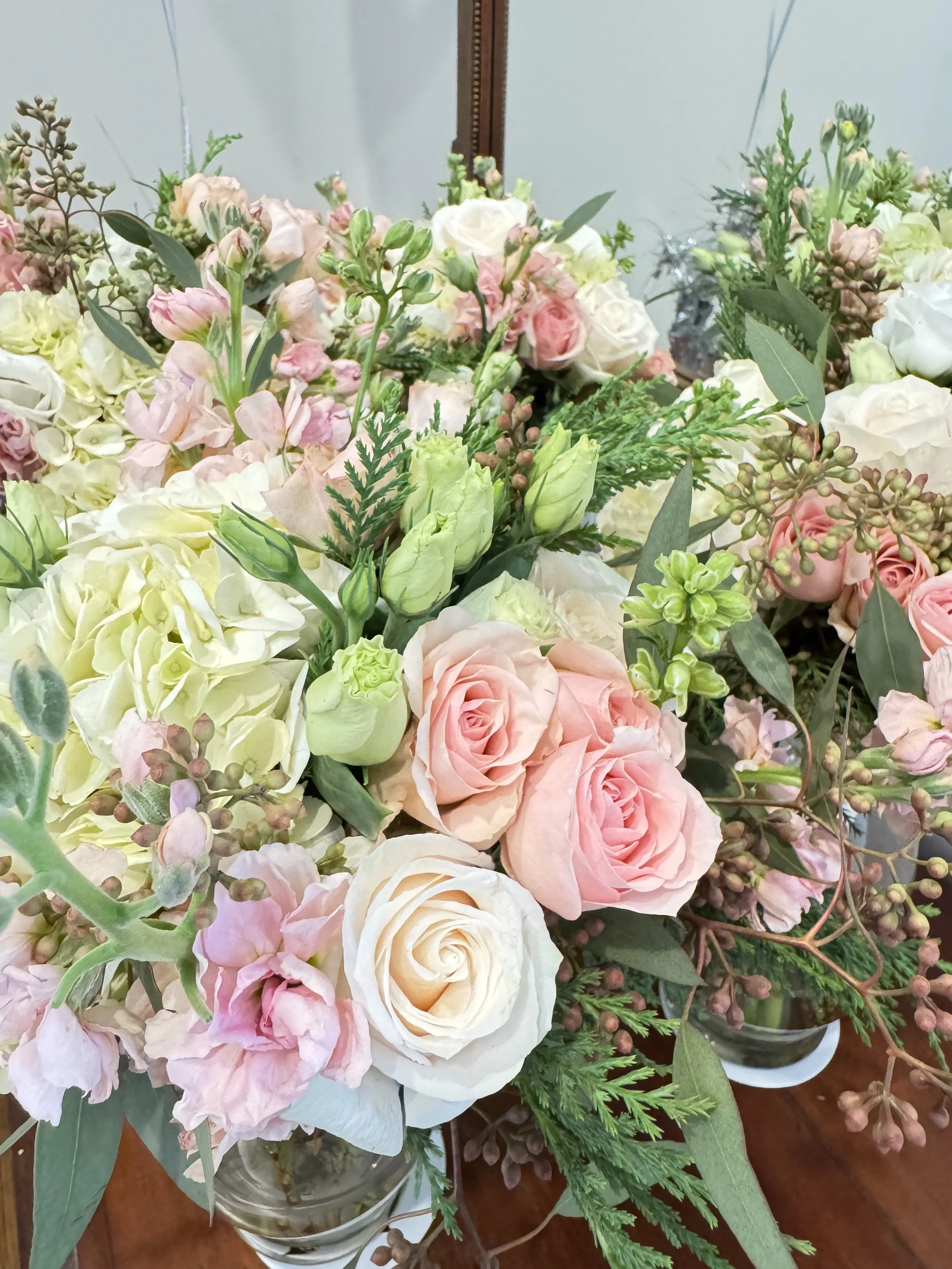 A vase of mixed pastel-colored flowers, including roses, peonies, and greenery, on a wooden surface.