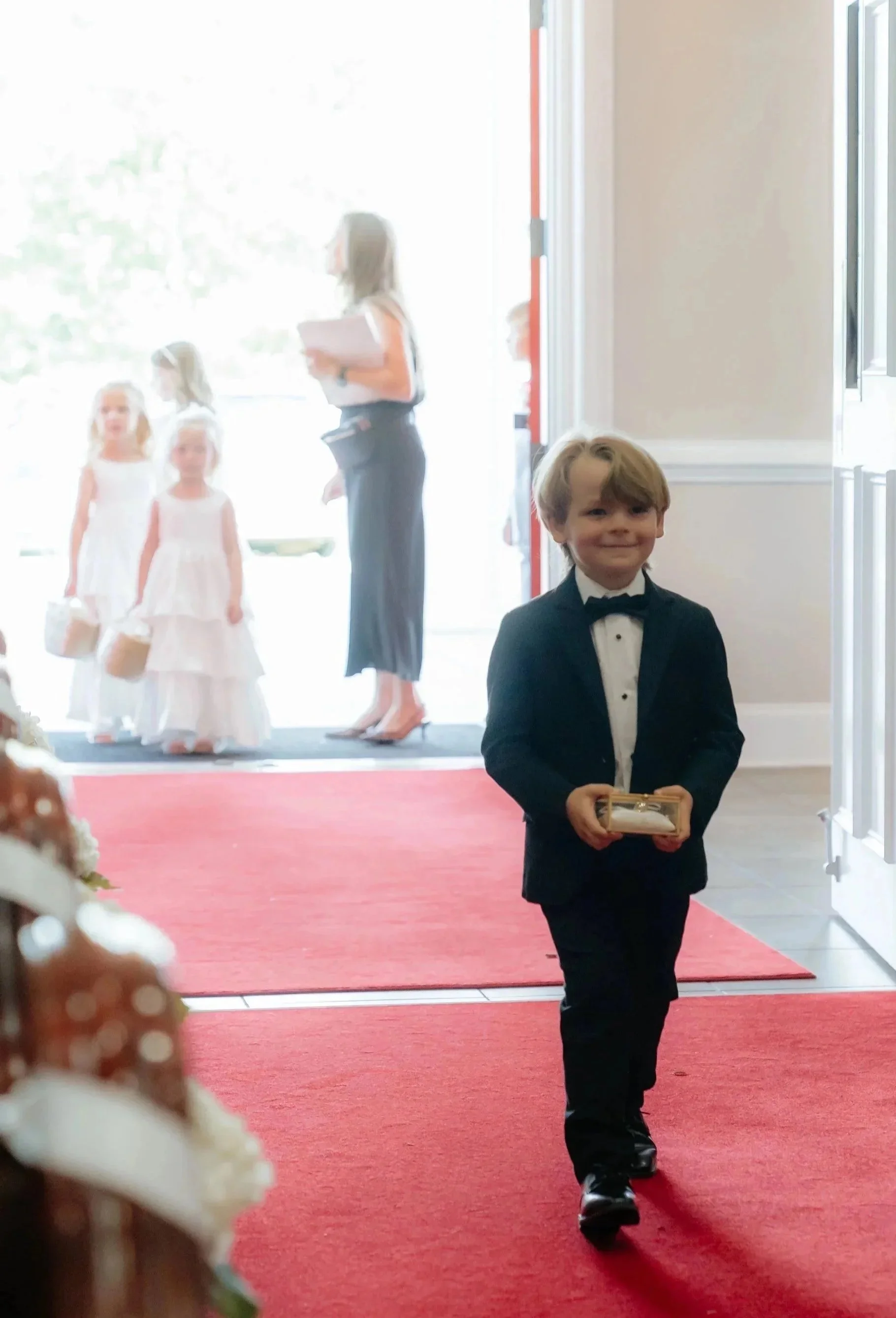 A young boy dressed in a tuxedo holding a small box, walking on a red carpet inside a building, with a woman and children dressed in white dresses in the background, near an open bright doorway.