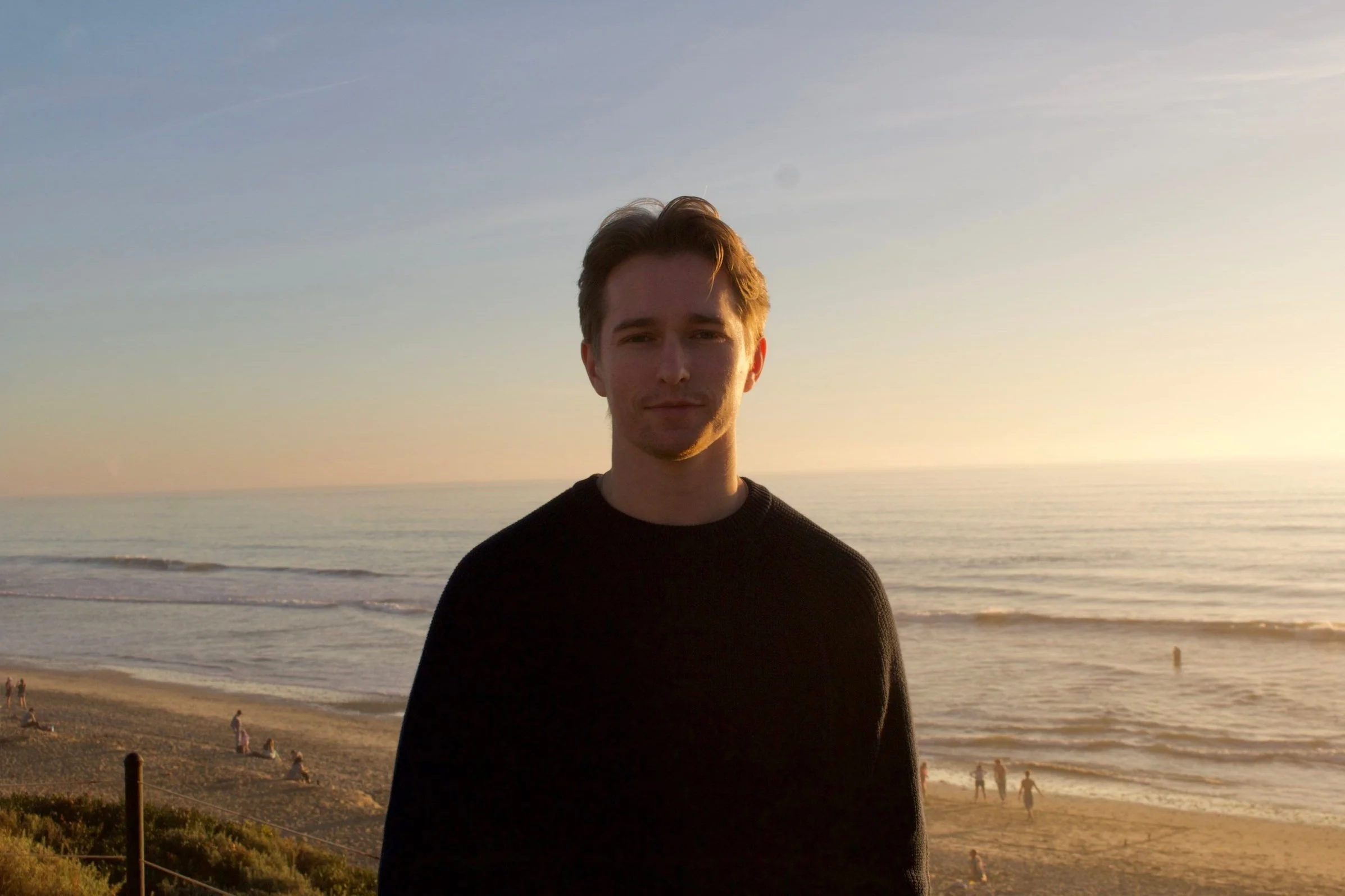 A young man with light brown hair standing on a beach at sunset, with the ocean and a few people in the background.