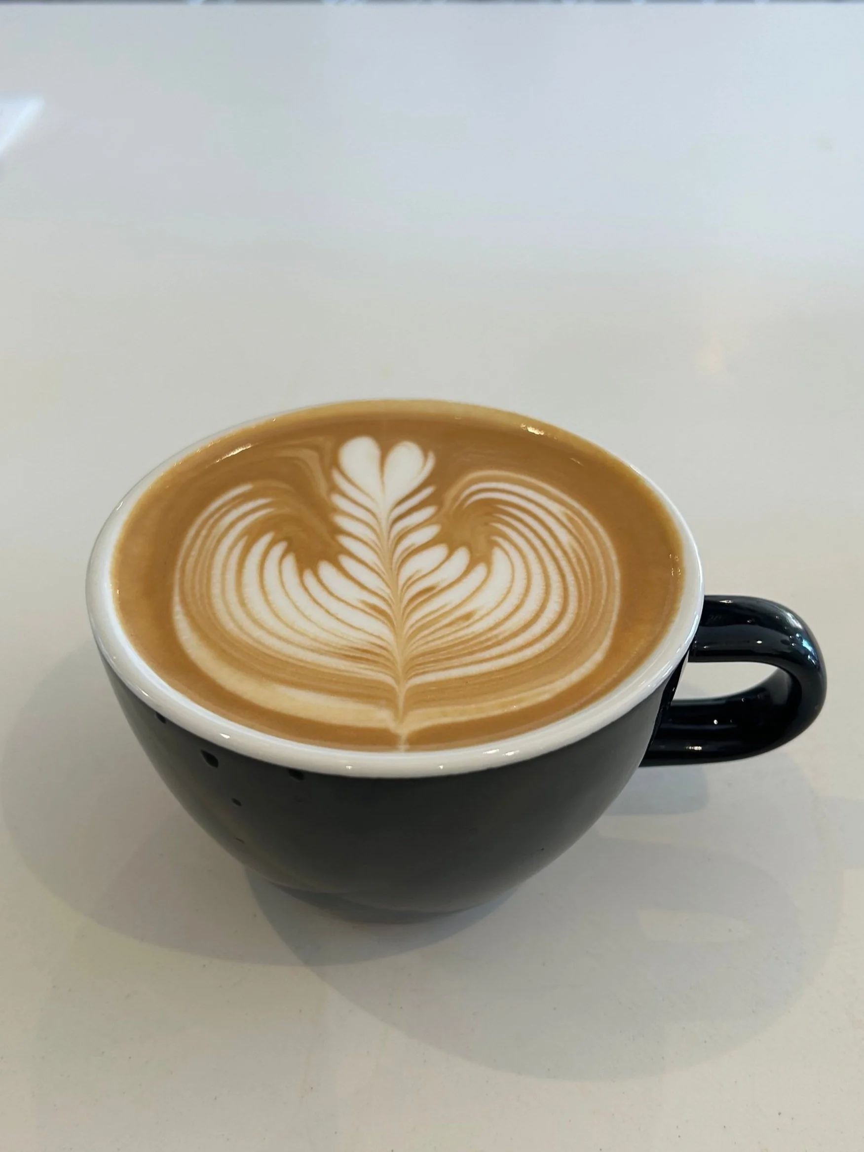 Black ceramic cup of latte with intricate white latte art heart and leaf design on top, on a white surface.