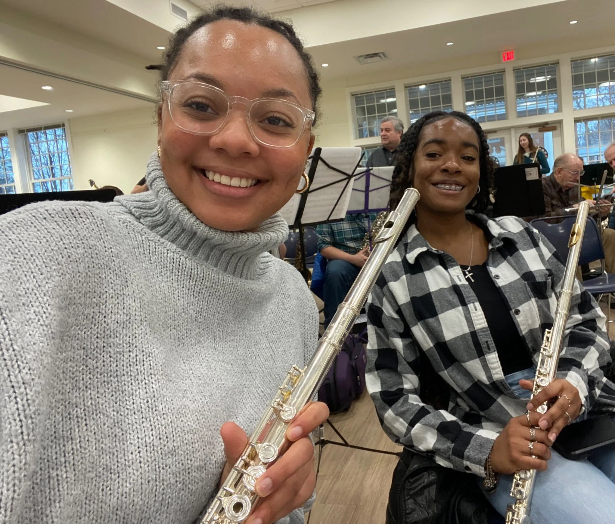 Two people holding flutes in a music rehearsal room, smiling at the camera. They are seated with other musicians in the background.