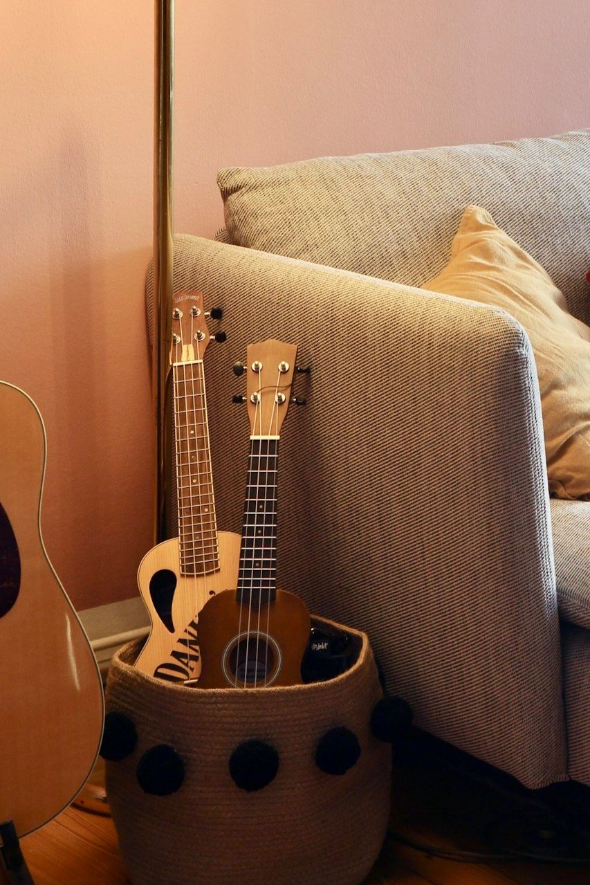 Two ukuleles in a woven basket next to a gray sofa and acoustic guitar, with a beige wall and pillow in the background.