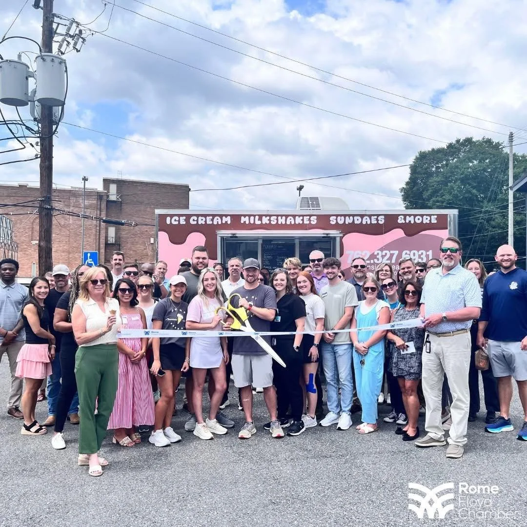 Group of people in front of a food trailer during daytime, celebrating a ribbon-cutting event with scissors and a ribbon, under cloudy sky.