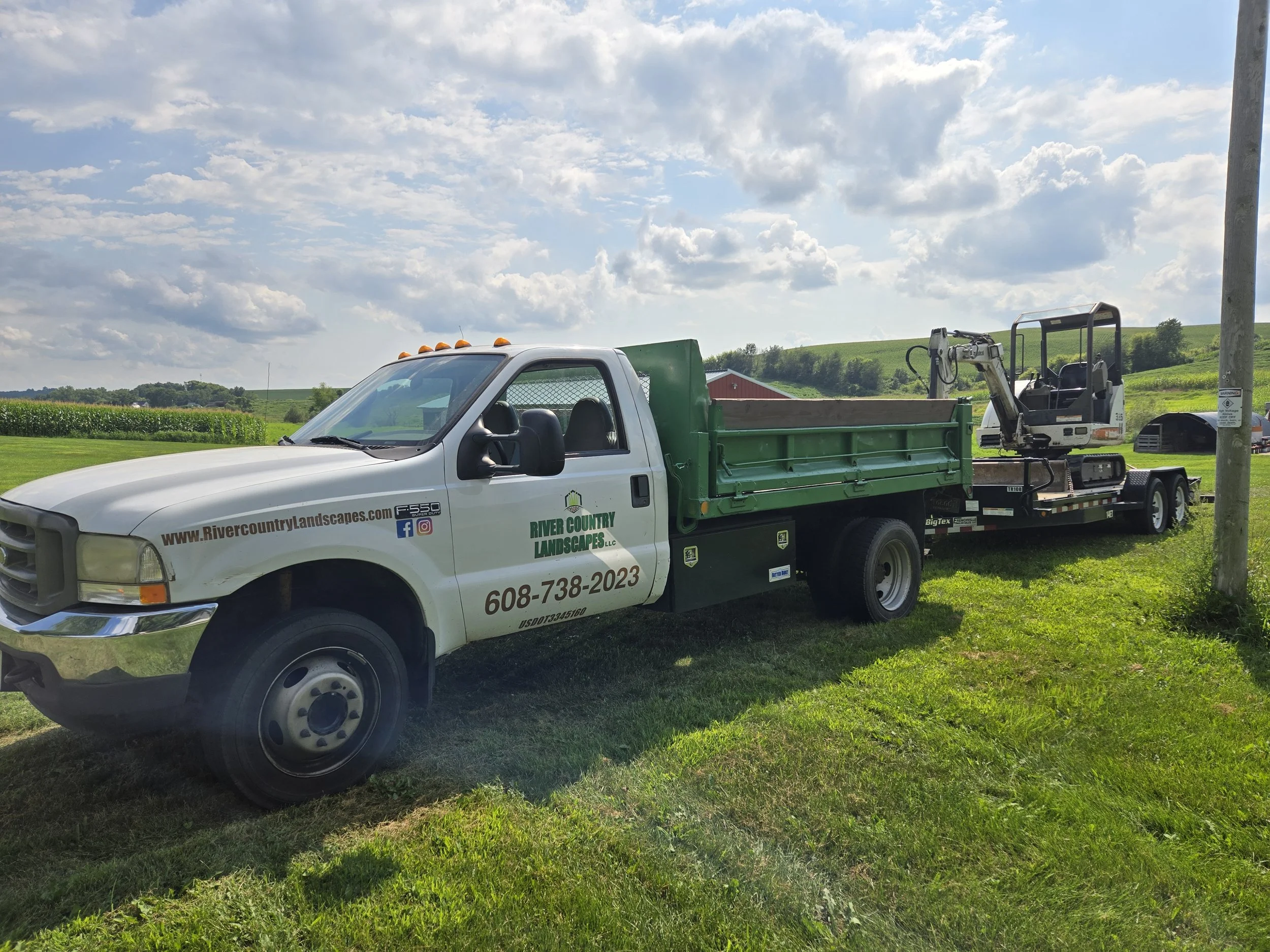 A white and green landscaping truck with equipment on a grassy field under a partly cloudy sky.