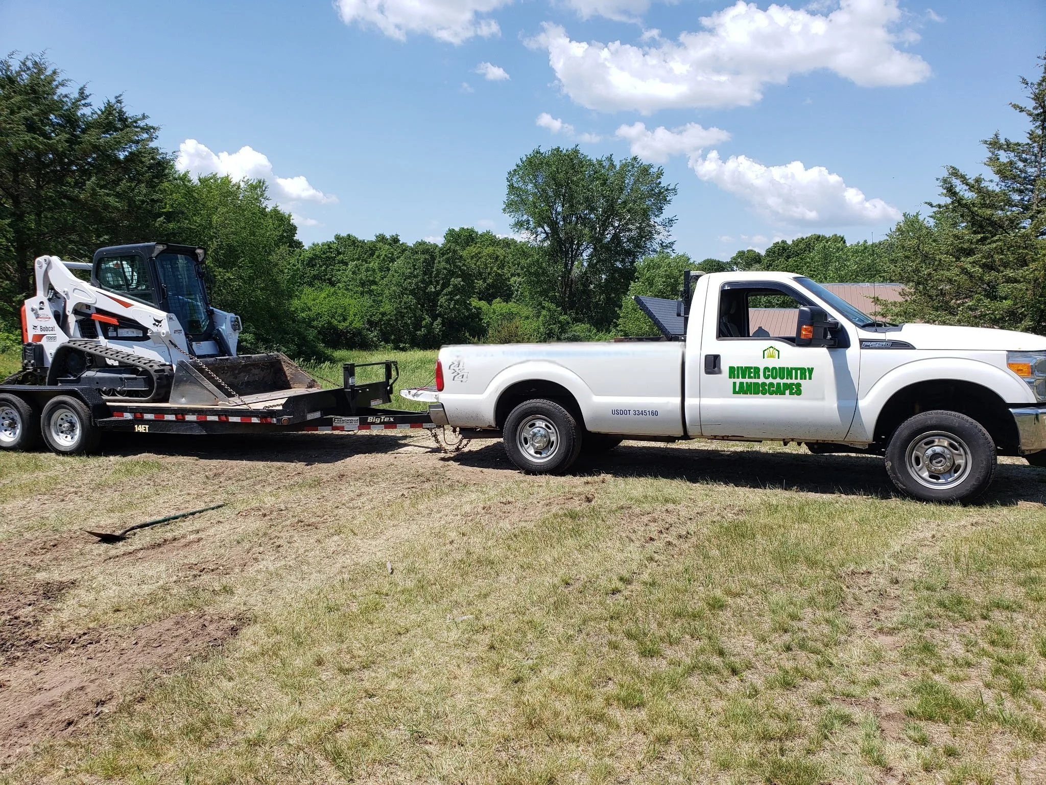 A white pickup truck with 'River Country Landscapes' written on the side, towing a flatbed trailer with a Bobcat skid steer loader.