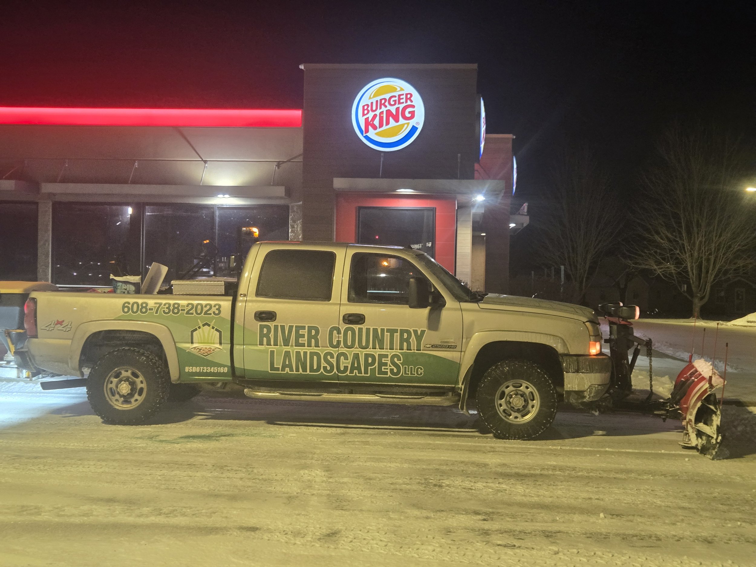 A yellow pickup truck with green lettering for River Country Landscapes is parked outside a Burger King at night, with snow on the ground and a snow plow attached to the front of the truck.